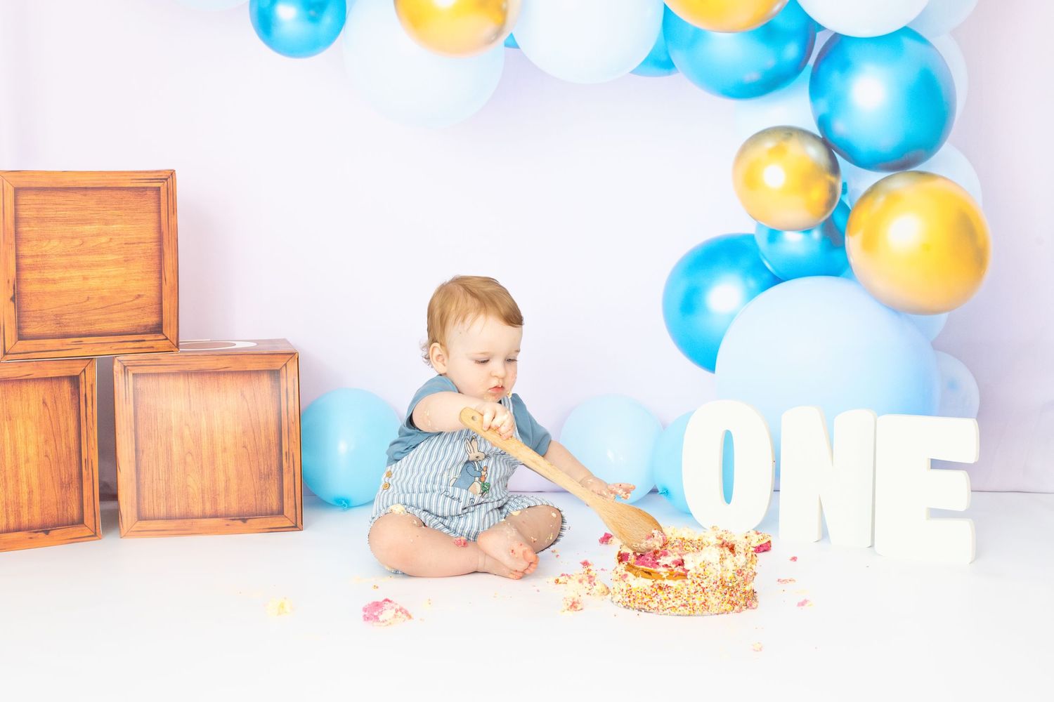 Festive first birthday scene with blue and gold balloons and white decorative elements.