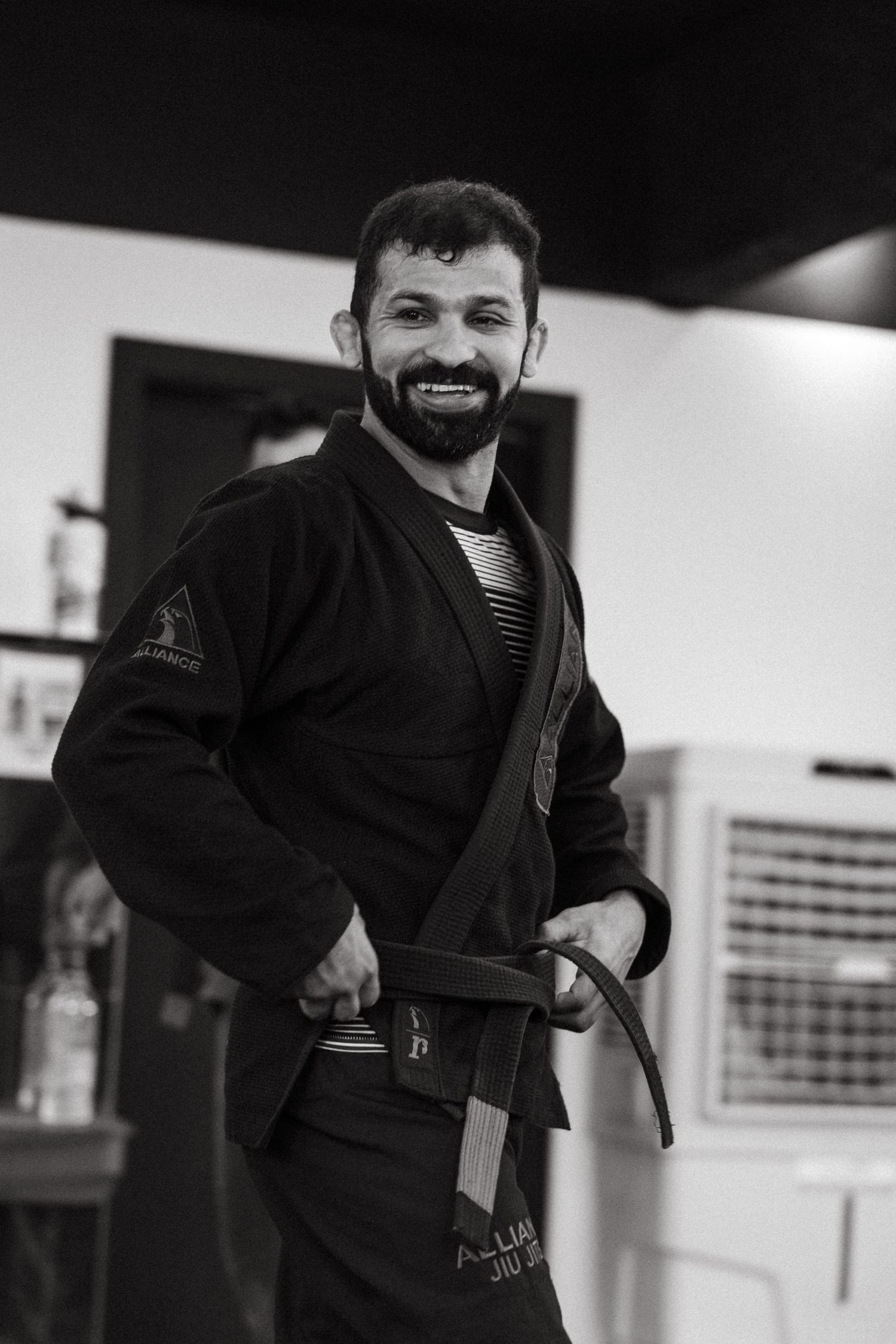 Black and white photo of a smiling bearded man (Bruno Malfacine) in a black jiu-jitsu gi with a black belt.