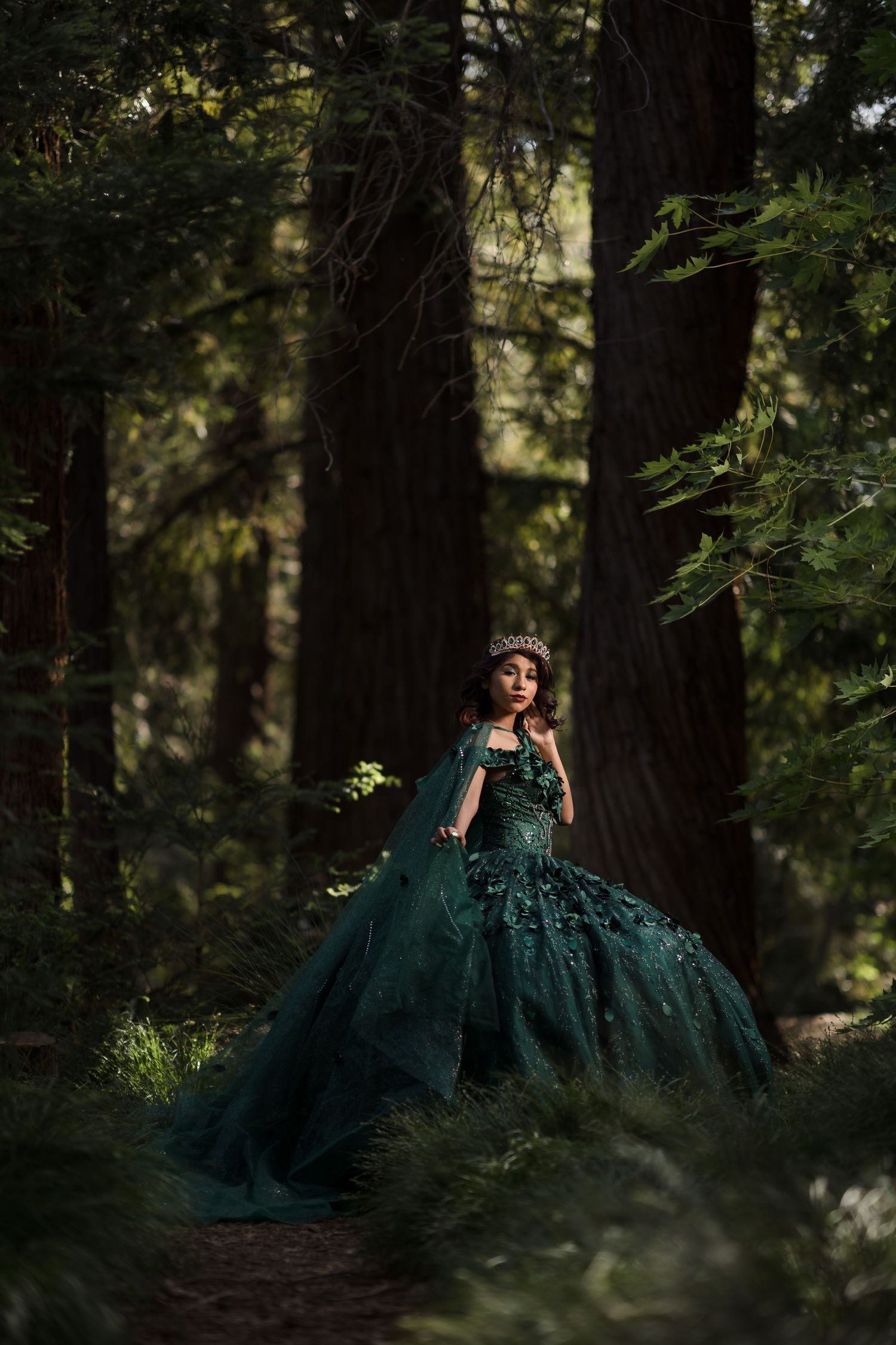 Woman in emerald green ballgown holding lantern in enchanted forest setting.