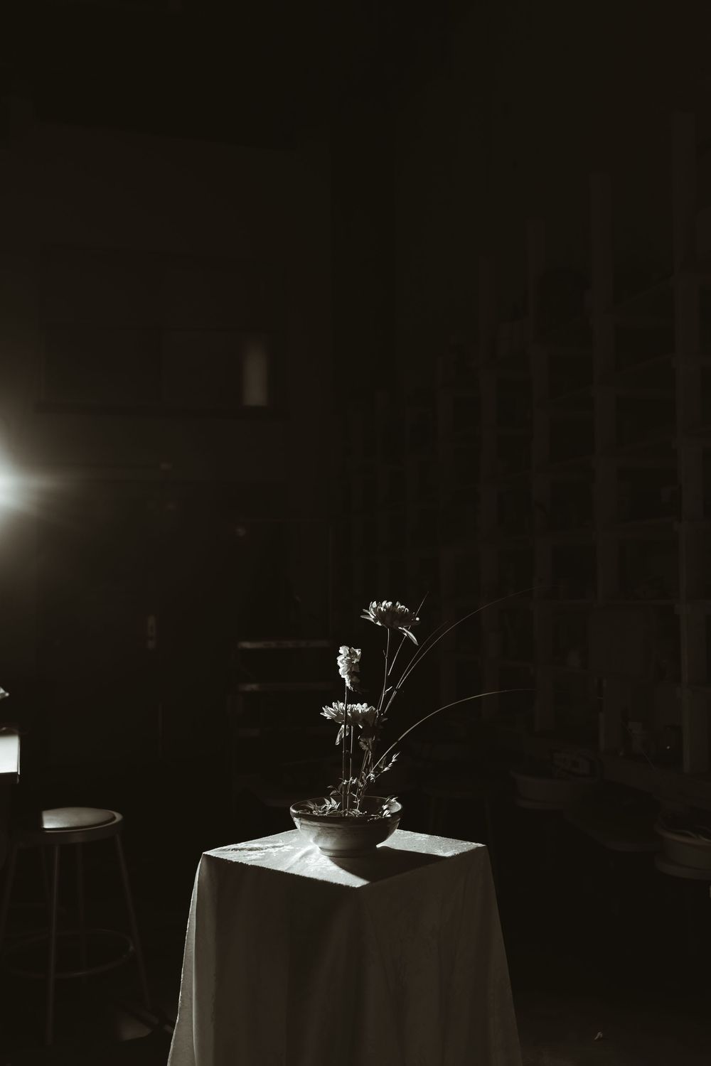Minimalist ikebana floral arrangement in a dark bowl spotlit on a white draped table in a dim room.