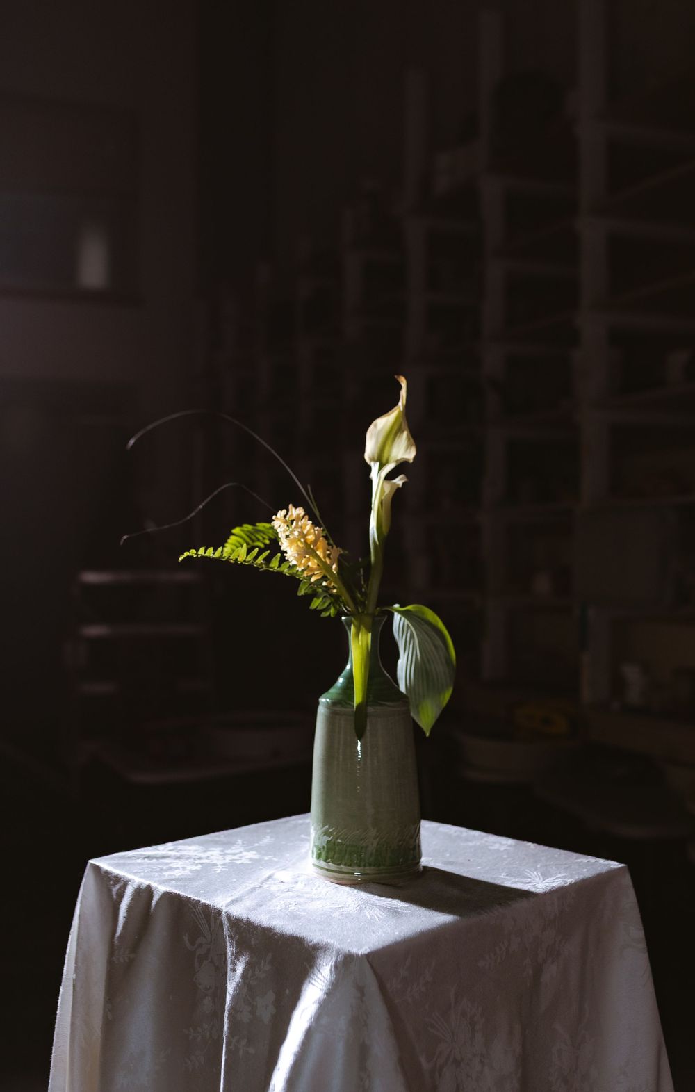 A white calla lily arrangement in a green ceramic vase on a white tablecloth, dramatically lit against a dark background.