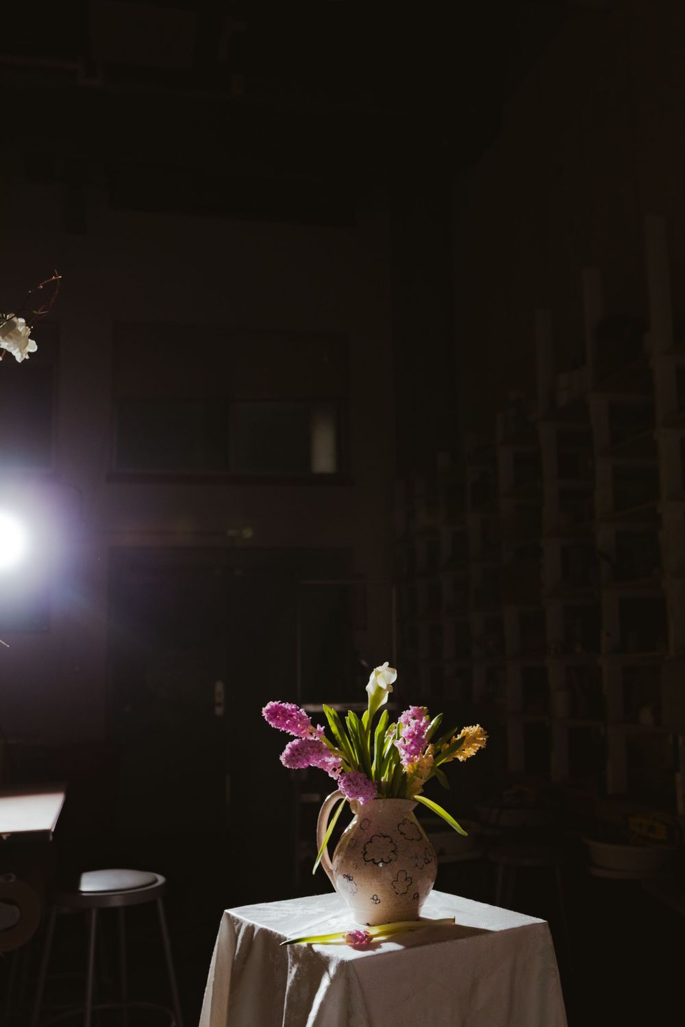 Pink and white tulips in a ceramic vase on a white draped table, dramatically lit in a dark room.