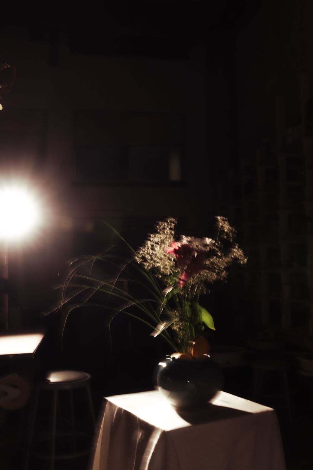 Dark moody floral arrangement in black vase on white draped table illuminated by bright spotlight.