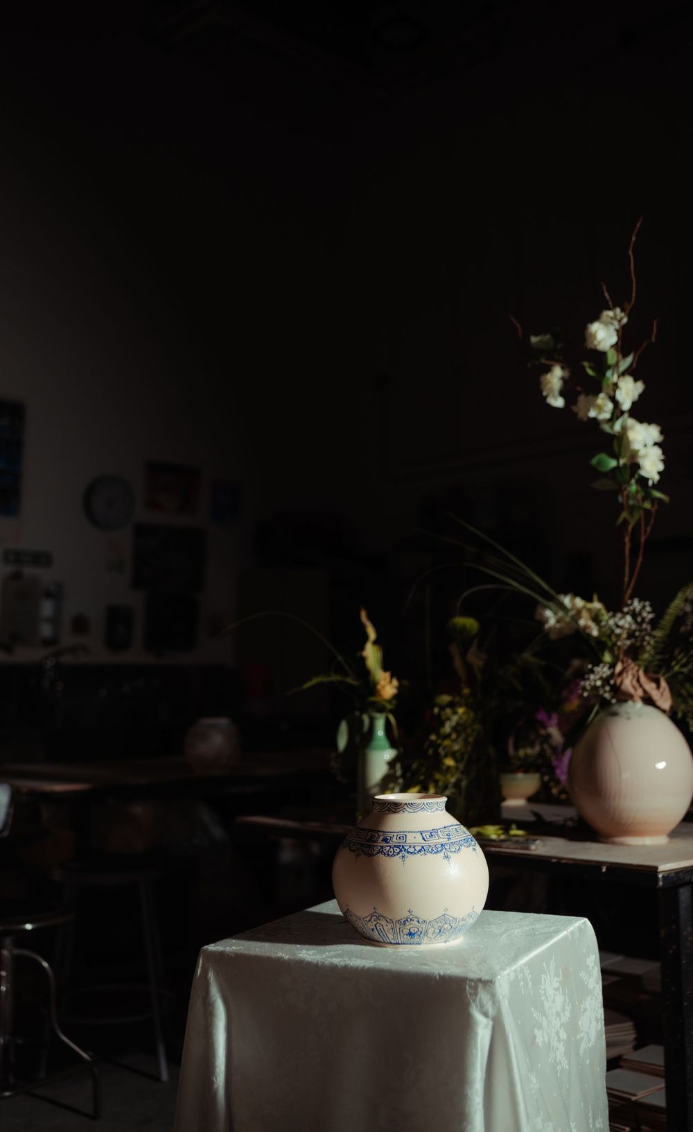 White ceramic vase with blue floral pattern on draped table in dark moody interior with flowers in background.