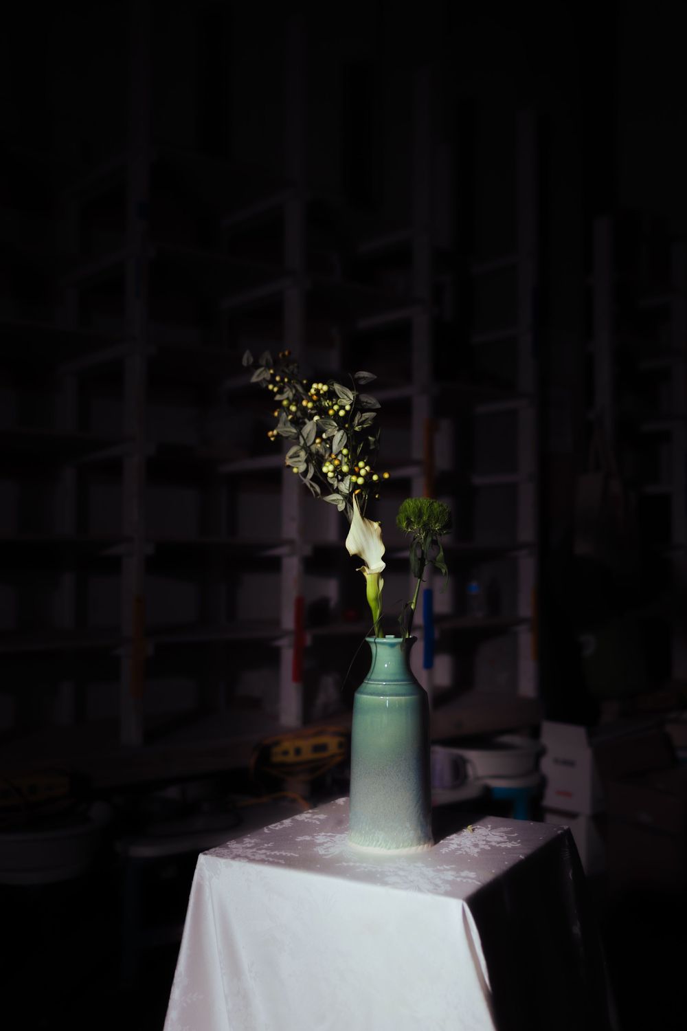 A ceramic vase with white calla lily and yellow berry branches on a white draped table in a dark room.
