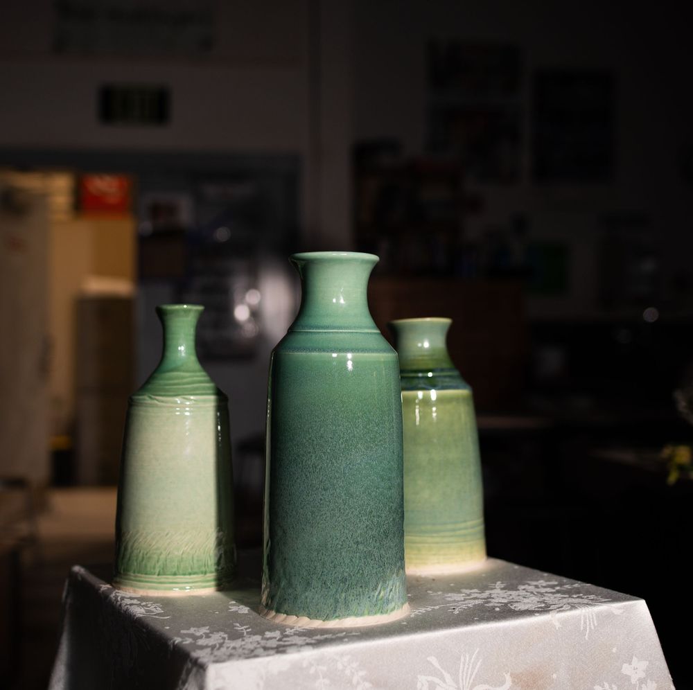 Three green ceramic vases with speckled glaze displayed on a white tablecloth in a dimly lit room.