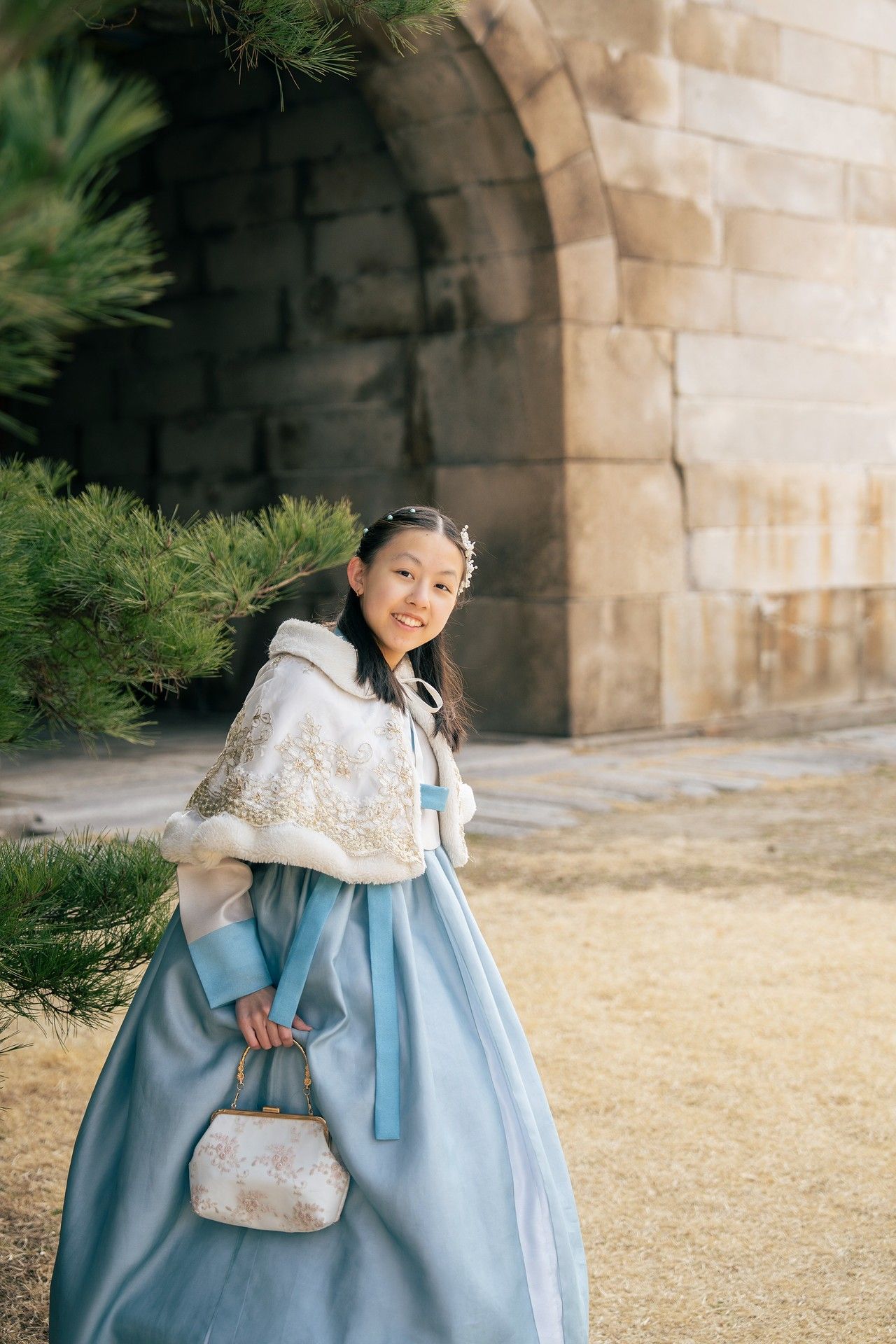 Young girl in traditional Korean hanbok dress with white cape and purse, posing near stone wall and pine tree.