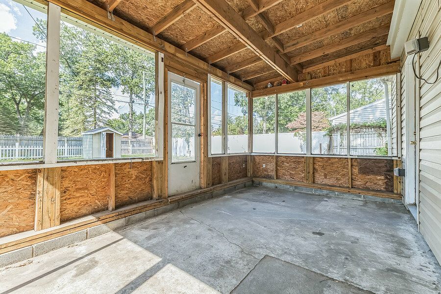 Unfinished screened porch with wood paneling, concrete floor, and large windows overlooking a tree-lined backyard.