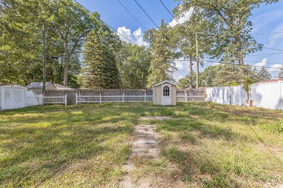 Spacious backyard with lush green grass, white vinyl fence, storage shed, and mature trees under a blue sky.