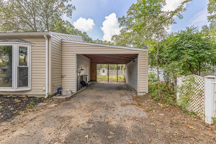 Covered carport with metal roof attached to a beige siding home surrounded by mature trees.