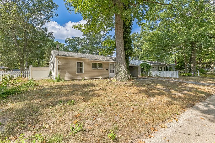 Single-story beige ranch home with large shade tree in front yard and white fence.