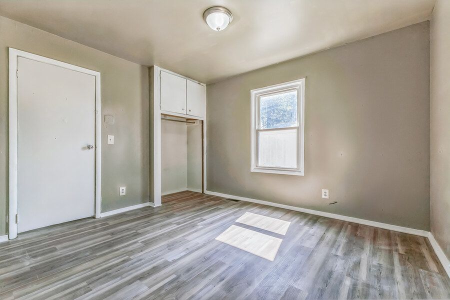 Bright empty bedroom with light gray walls, hardwood floors, white closet with upper cabinets, and a window.