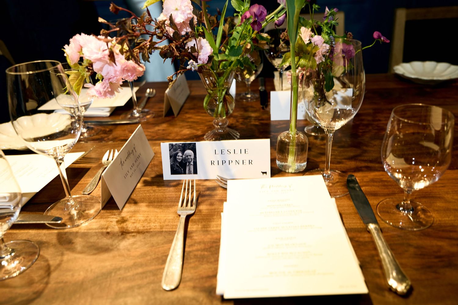Elegant wedding table setting with floral centerpieces, place cards, wine glasses, and silverware on a rustic wood table.