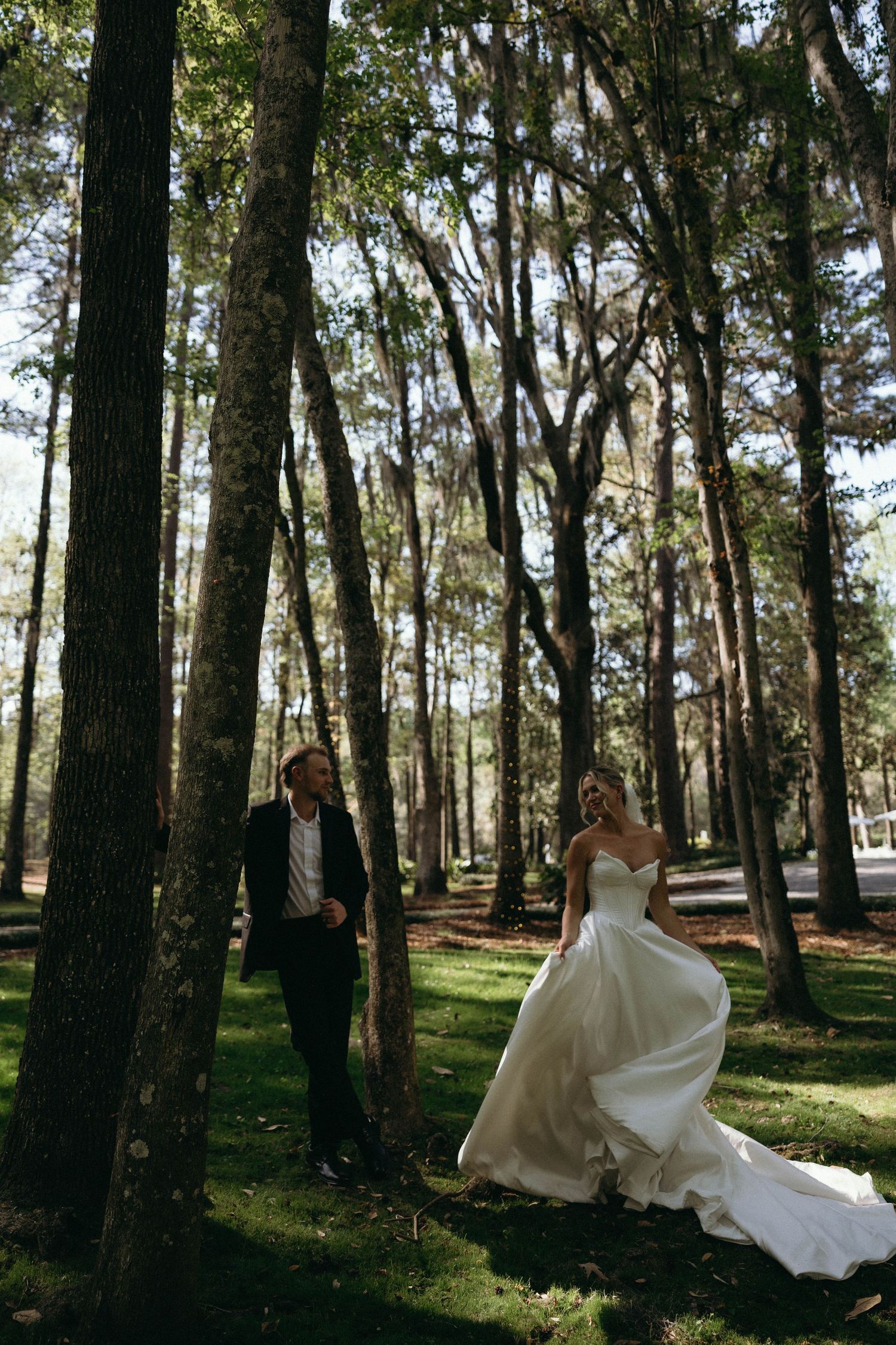 Bride in white gown and groom in black suit walk among tall trees in a sunlit forest during wedding photos.
