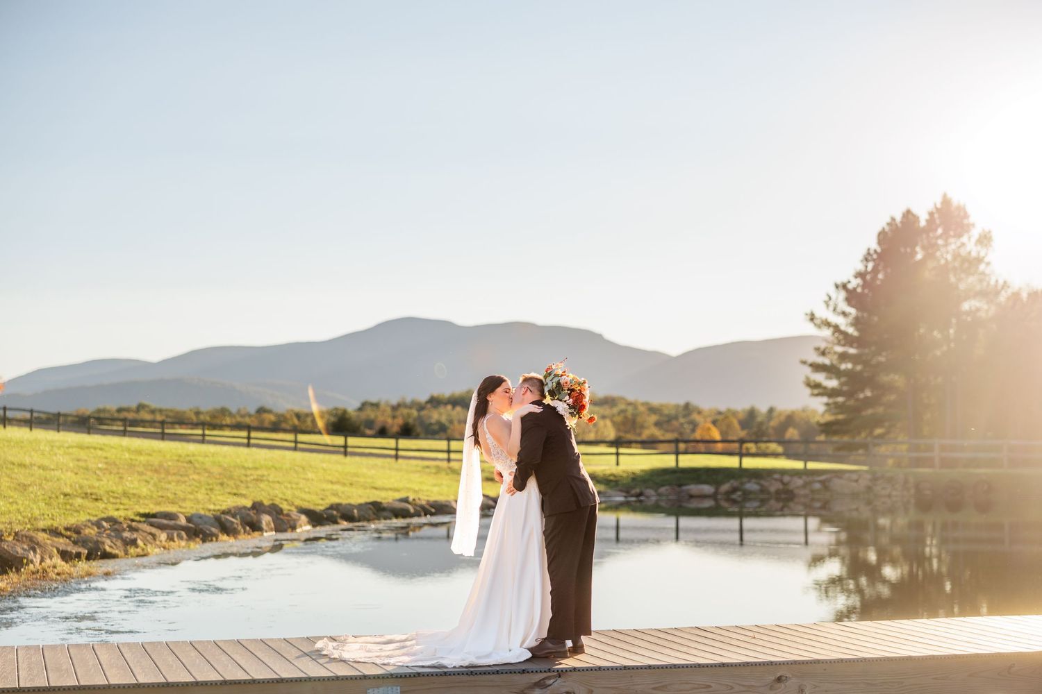 Bride and groom sharing a kiss on a wooden dock beside a pond with mountain views and golden sunlight.