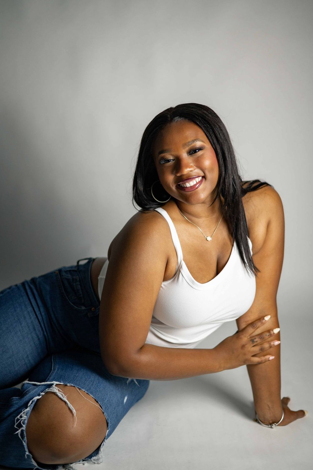 Smiling young Black woman in white tank top and ripped jeans sitting on floor in studio photo.