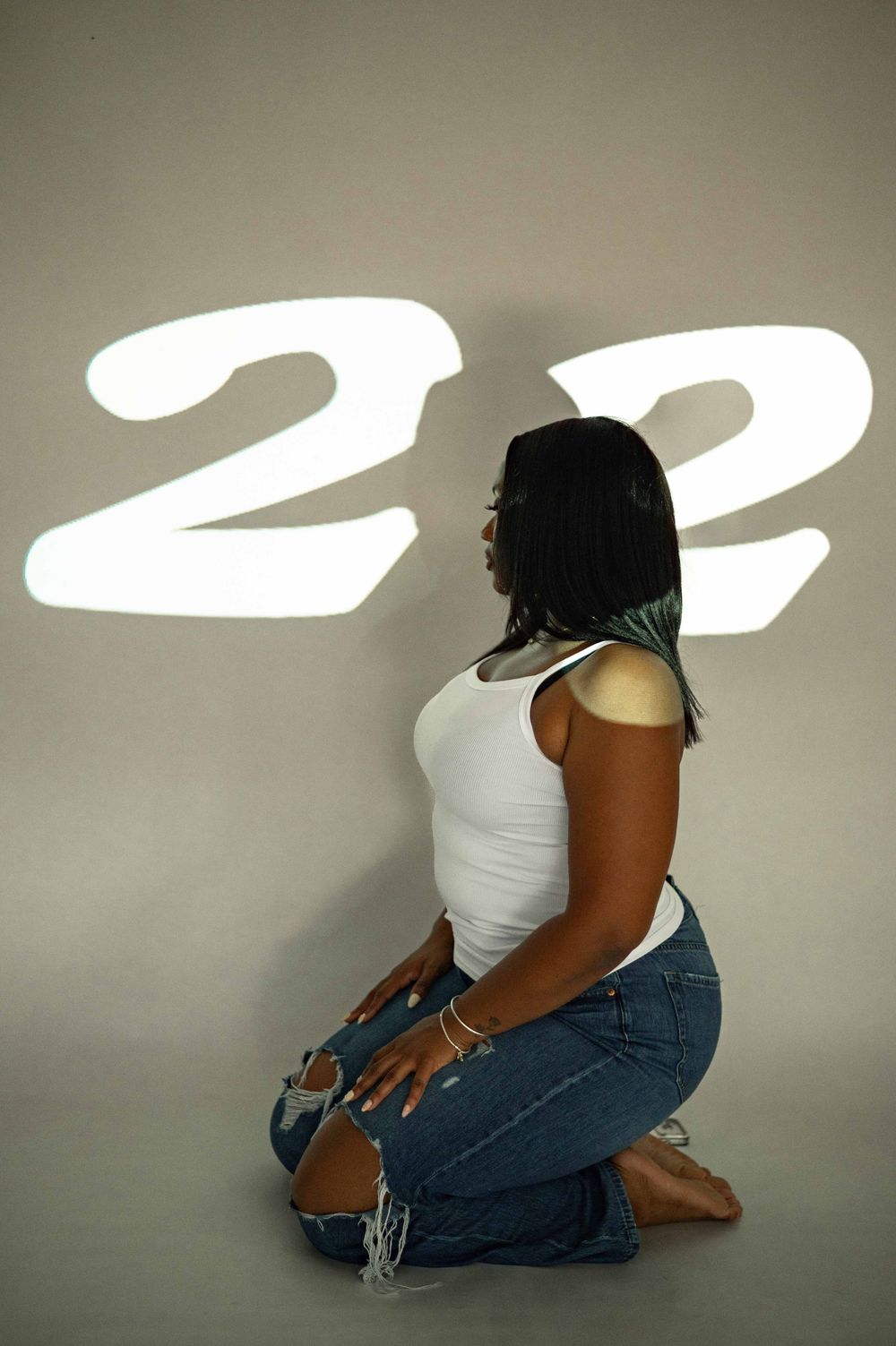 Young Black woman kneeling in white tank top and ripped jeans with number 22 projected on wall.