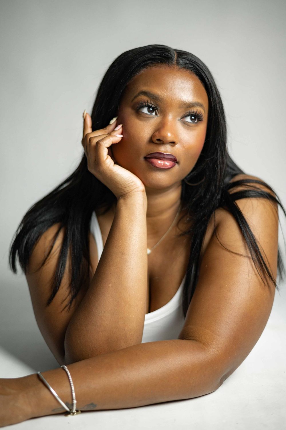 Young Black woman with long hair rests chin on hand, gazing upward in a white top against a gray background.