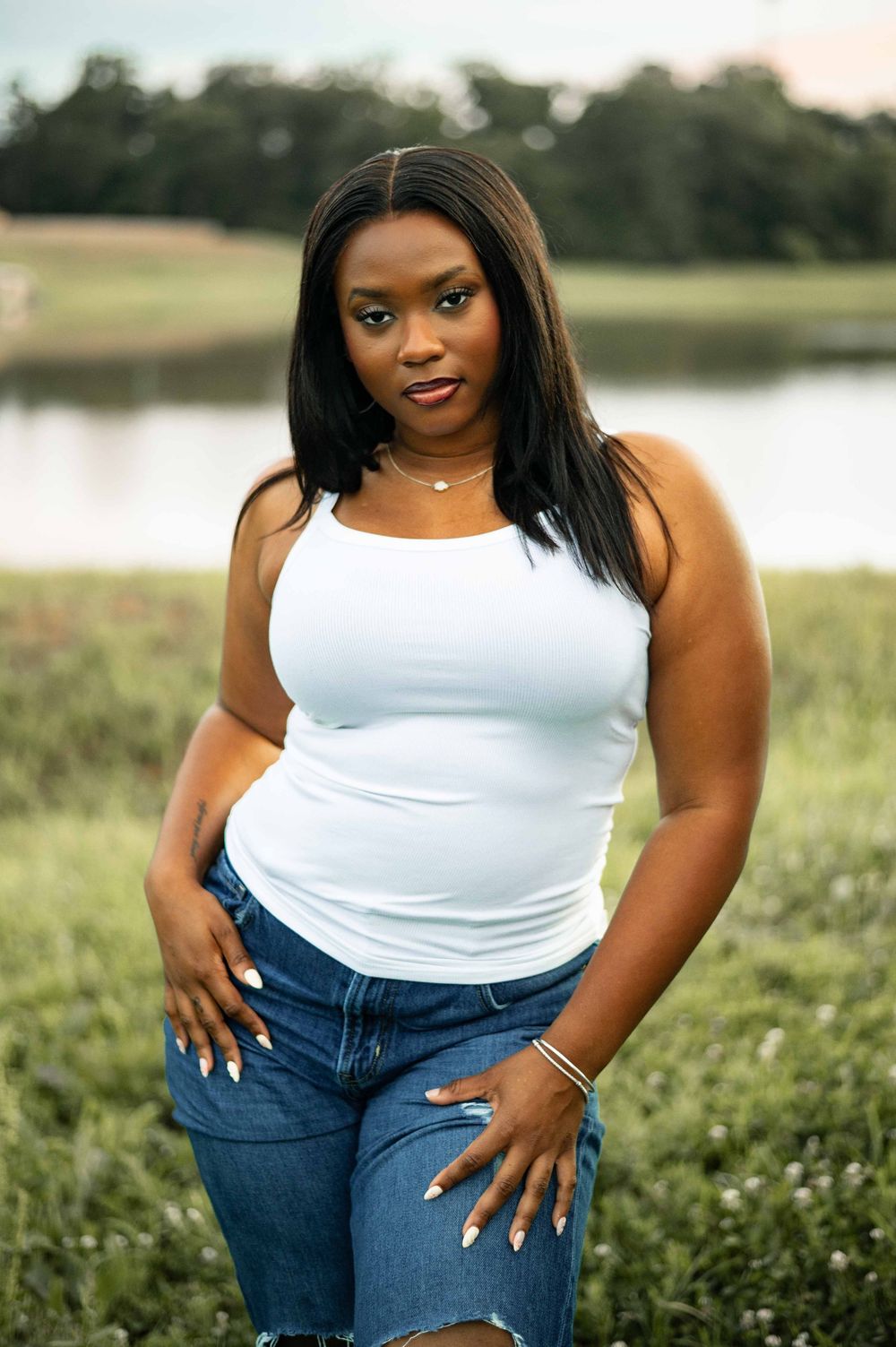 Young Black woman in white tank top and ripped jeans poses outdoors near a pond in a grassy field.
