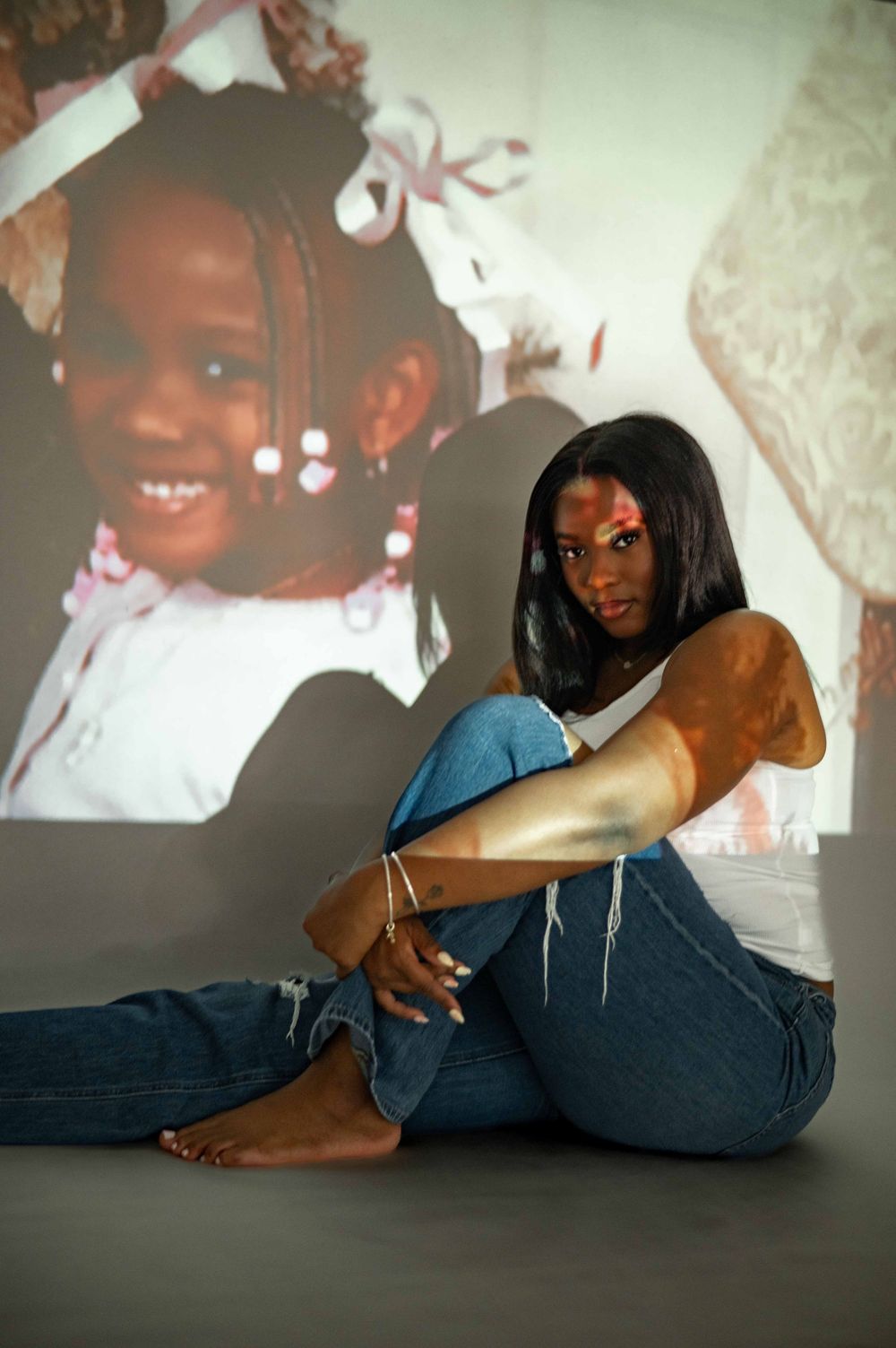 A young Black woman in ripped jeans sits barefoot against a wall with a projected childhood photo behind her.