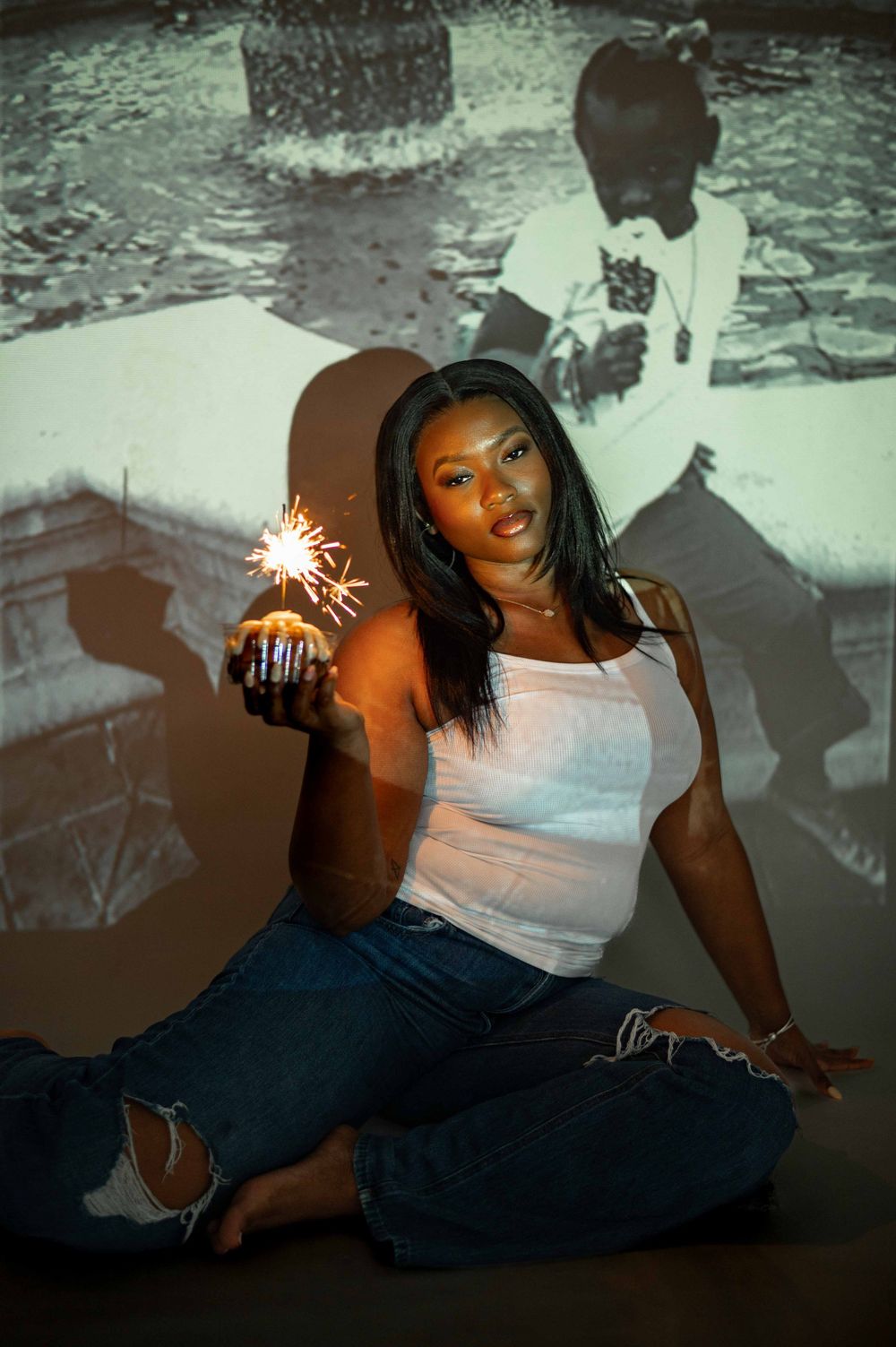 Woman in white tank and jeans holds sparkler cupcake before black and white projected photo.