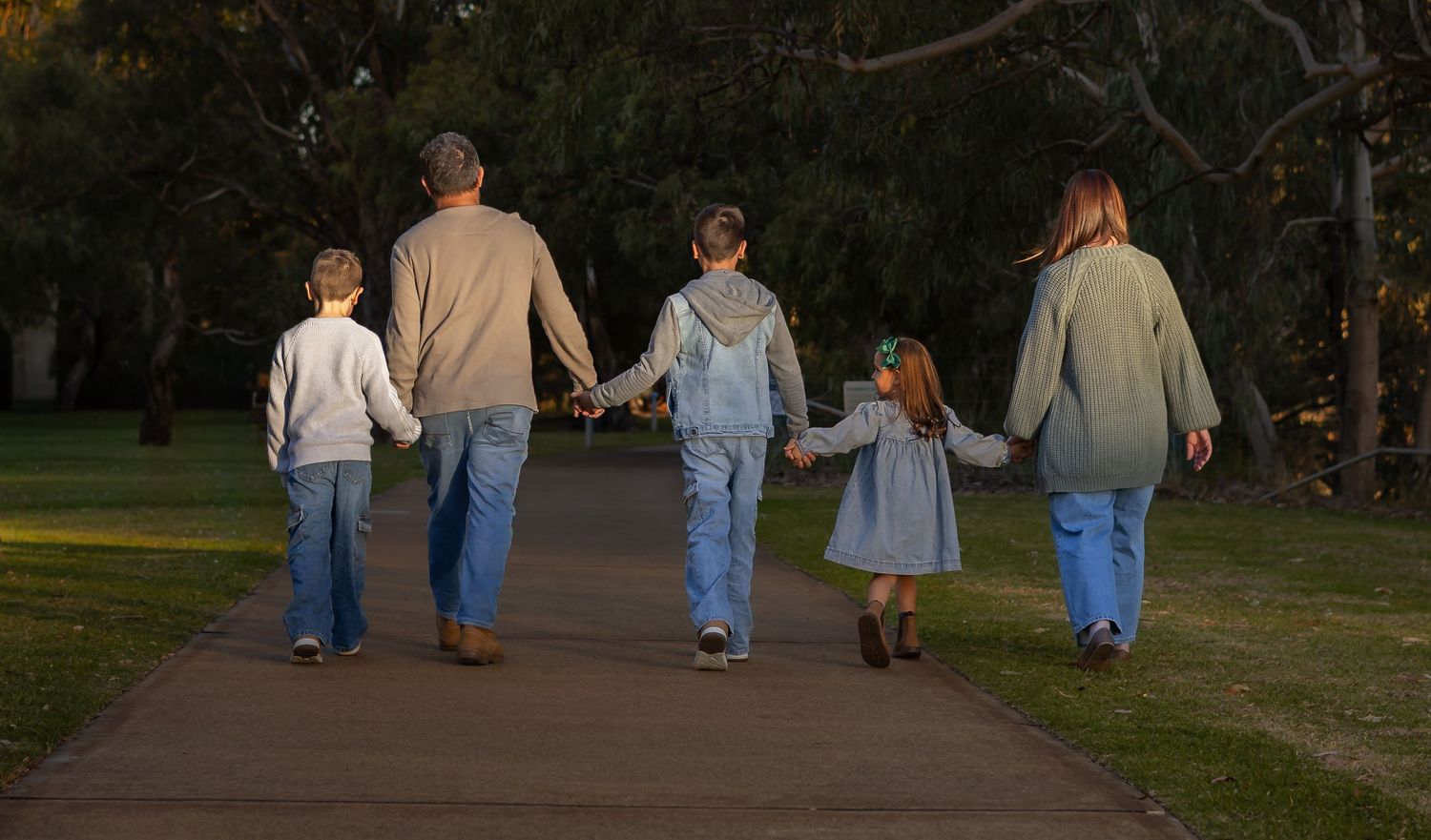 Family of five walking hand-in-hand down a park path surrounded by lush green trees at dusk.