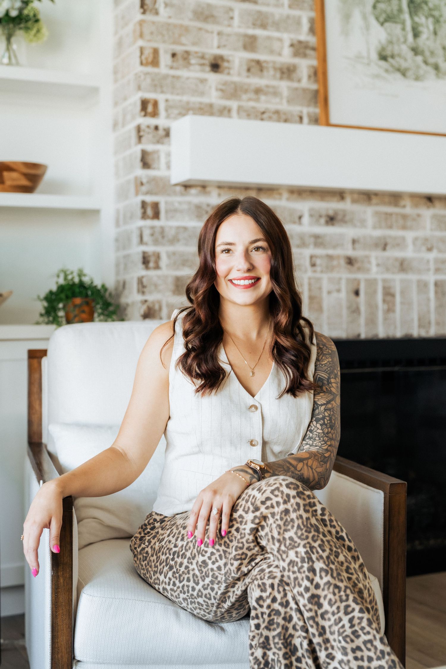 Smiling brunette woman in white top and leopard print pants seated in a chair beside a brick fireplace.