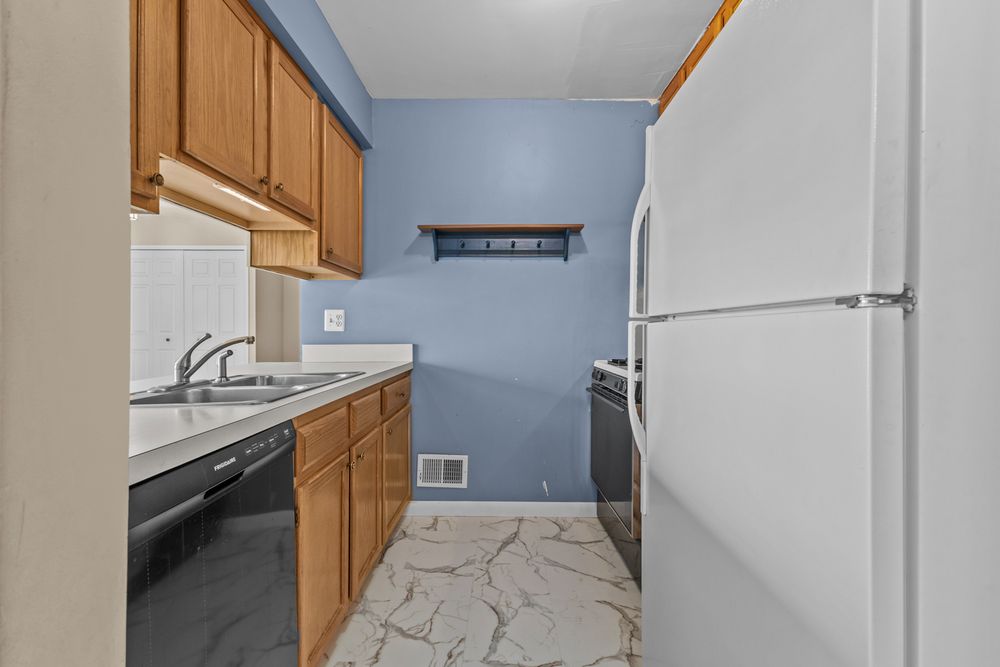 Compact kitchen with oak cabinets, black dishwasher, white fridge, blue walls, and marble-look floors.