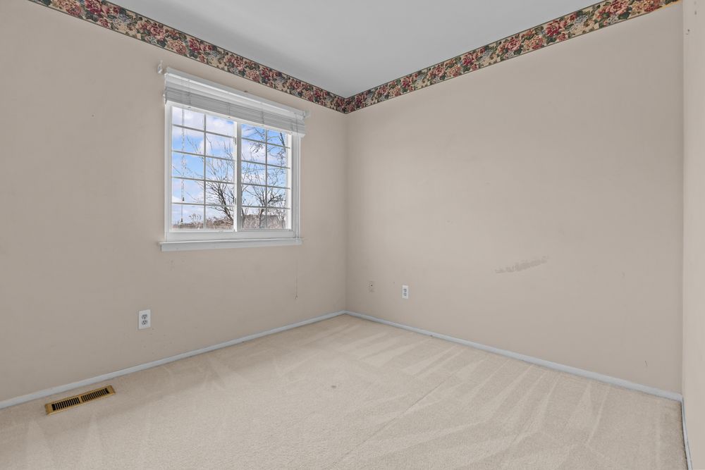 Empty bedroom with neutral carpet, floral wallpaper border, and bright window with natural light.