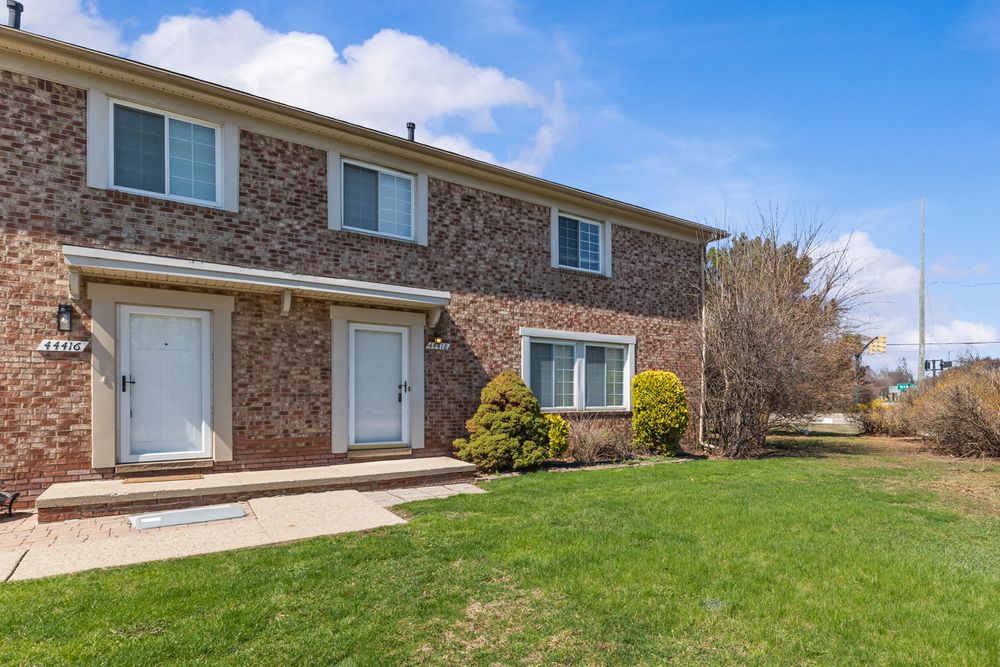Brick two-story townhouse with white entry doors, manicured lawn, and landscaped shrubs on a sunny day.