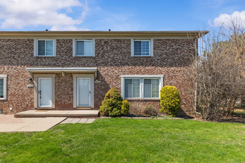 Two-story brick duplex with white entry doors, trimmed hedges, and green lawn in daylight.