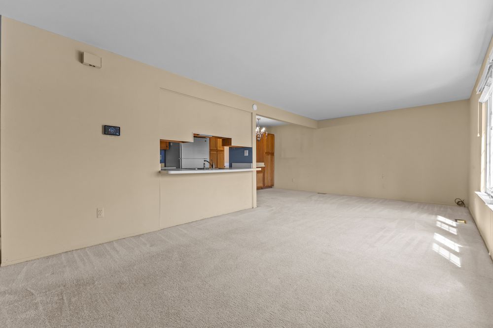 Open-concept living area with beige carpet, neutral walls, and view into kitchen with wood cabinets.