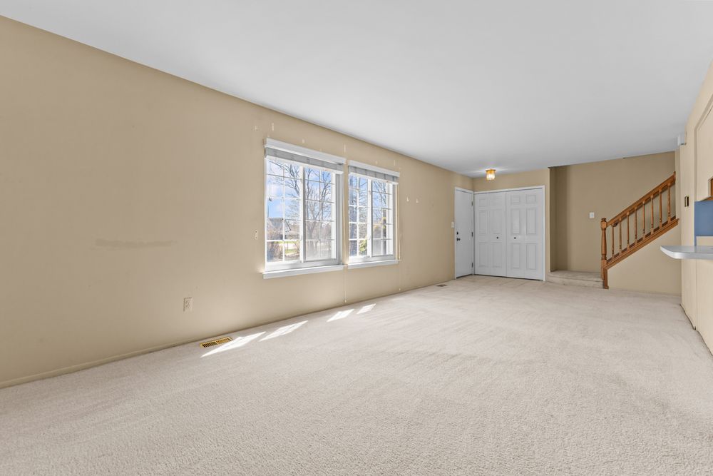 Bright empty living room with beige carpet, staircase, closet, and large front window.