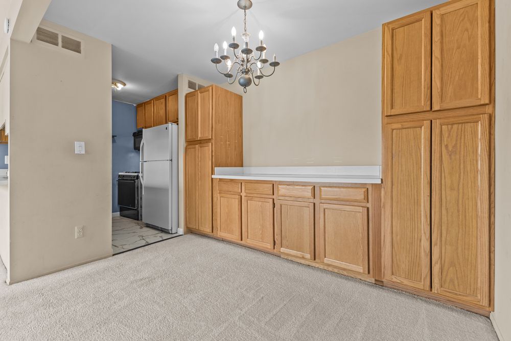 Kitchen area with oak cabinets, white refrigerator, beige carpet, and elegant chandelier overhead.