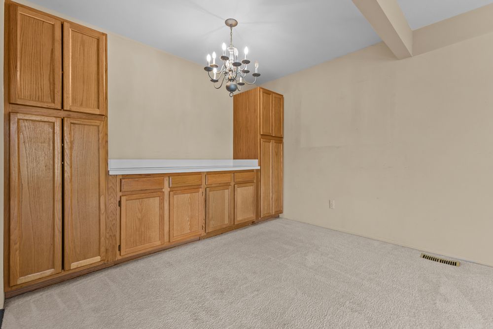 Spacious room with tall oak cabinets, built-in desk, beamed ceiling, chandelier, and neutral carpet.
