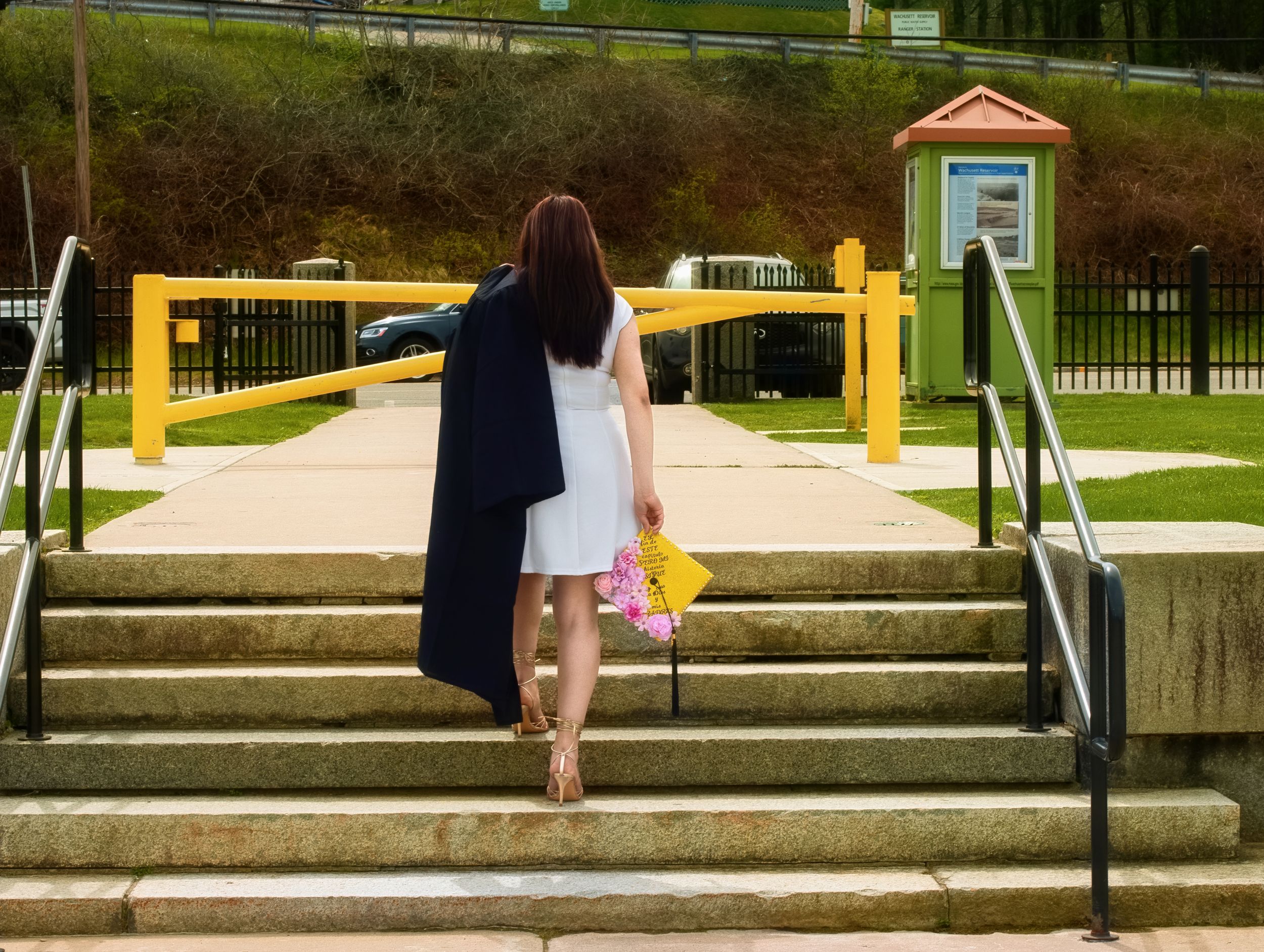 Graduate in black gown walks away carrying flowers up outdoor steps at a park.