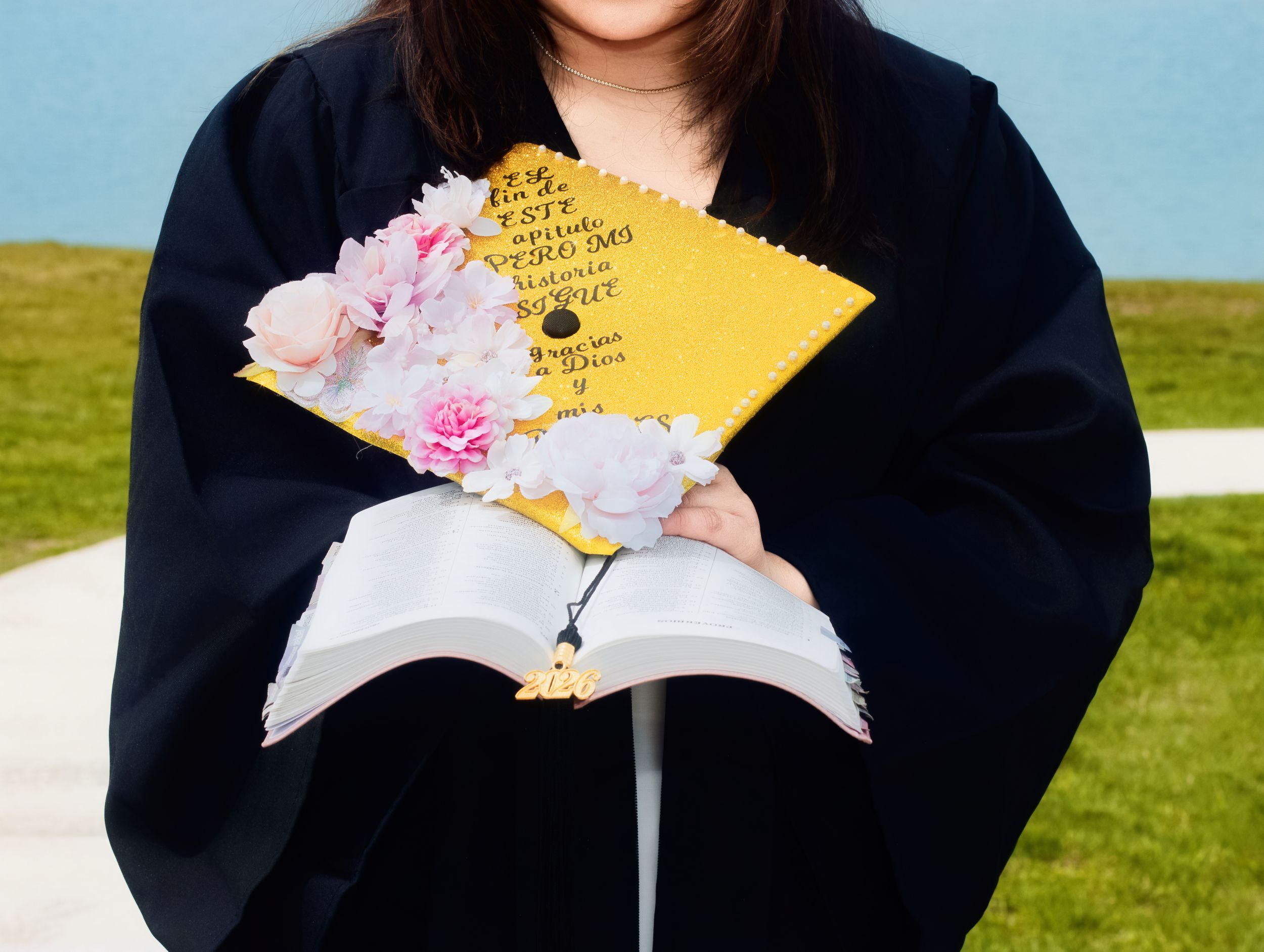 Graduate in black gown holds open book and decorated yellow mortarboard cap with pink flowers outdoors.