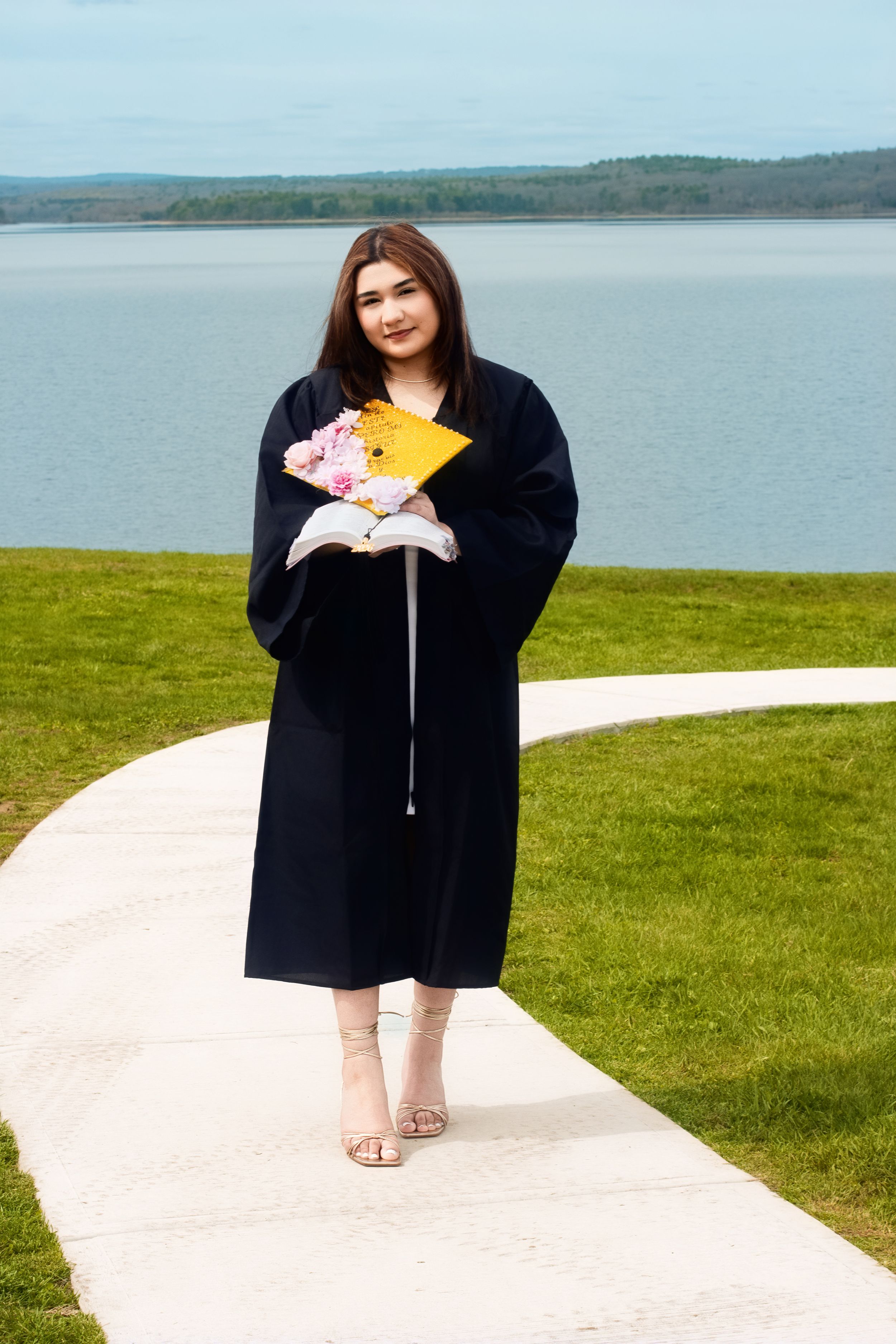 Young woman in black graduation gown holding yellow cap and flowers, standing on pathway by a lake.