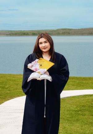 Graduate in black gown holding gold cap and flowers, posing by a scenic lake on a sunny day.