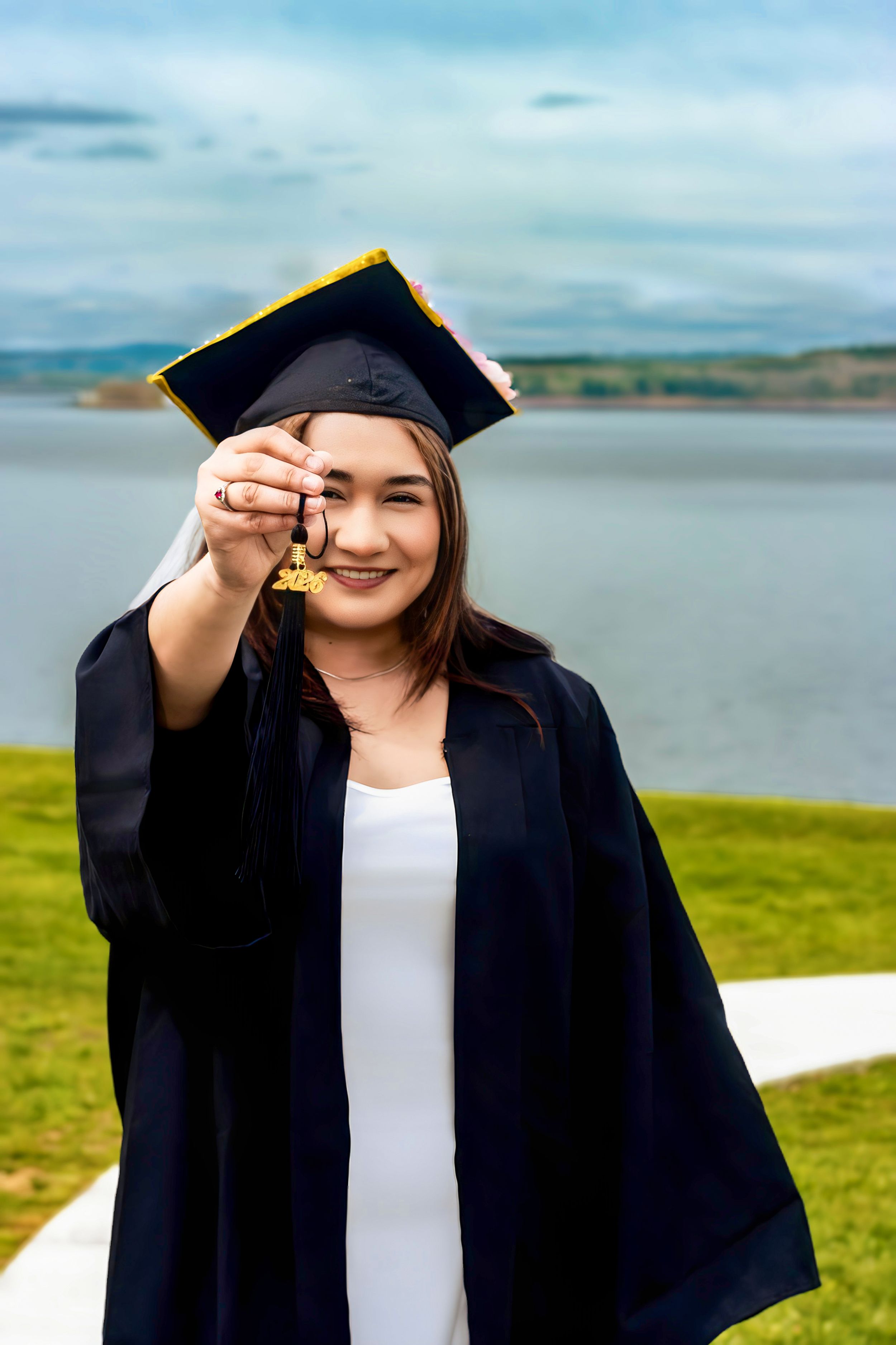 Smiling female graduate in black cap and gown holds tassel toward camera with lake and green lawn in background.