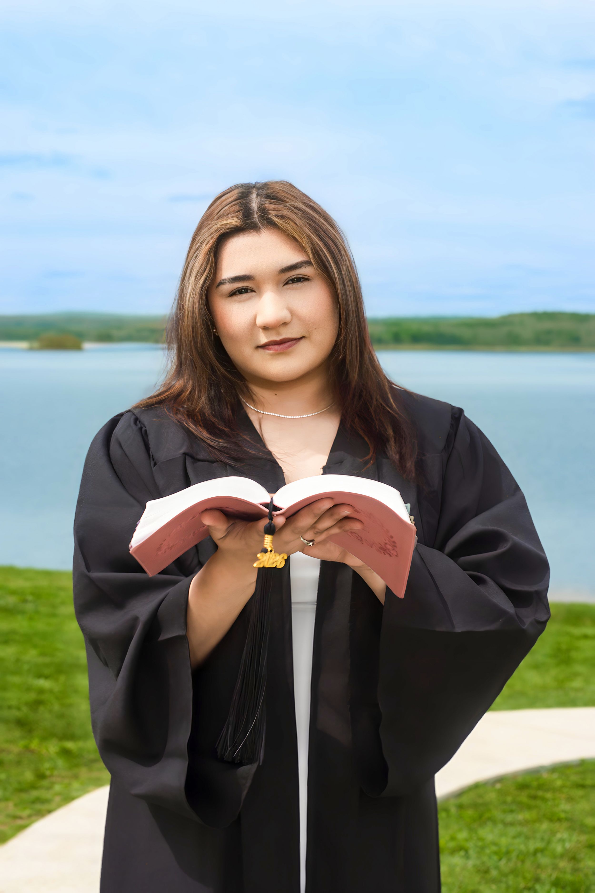 Young woman in black graduation gown holding open book near lakeside, smiling confidently outdoors.