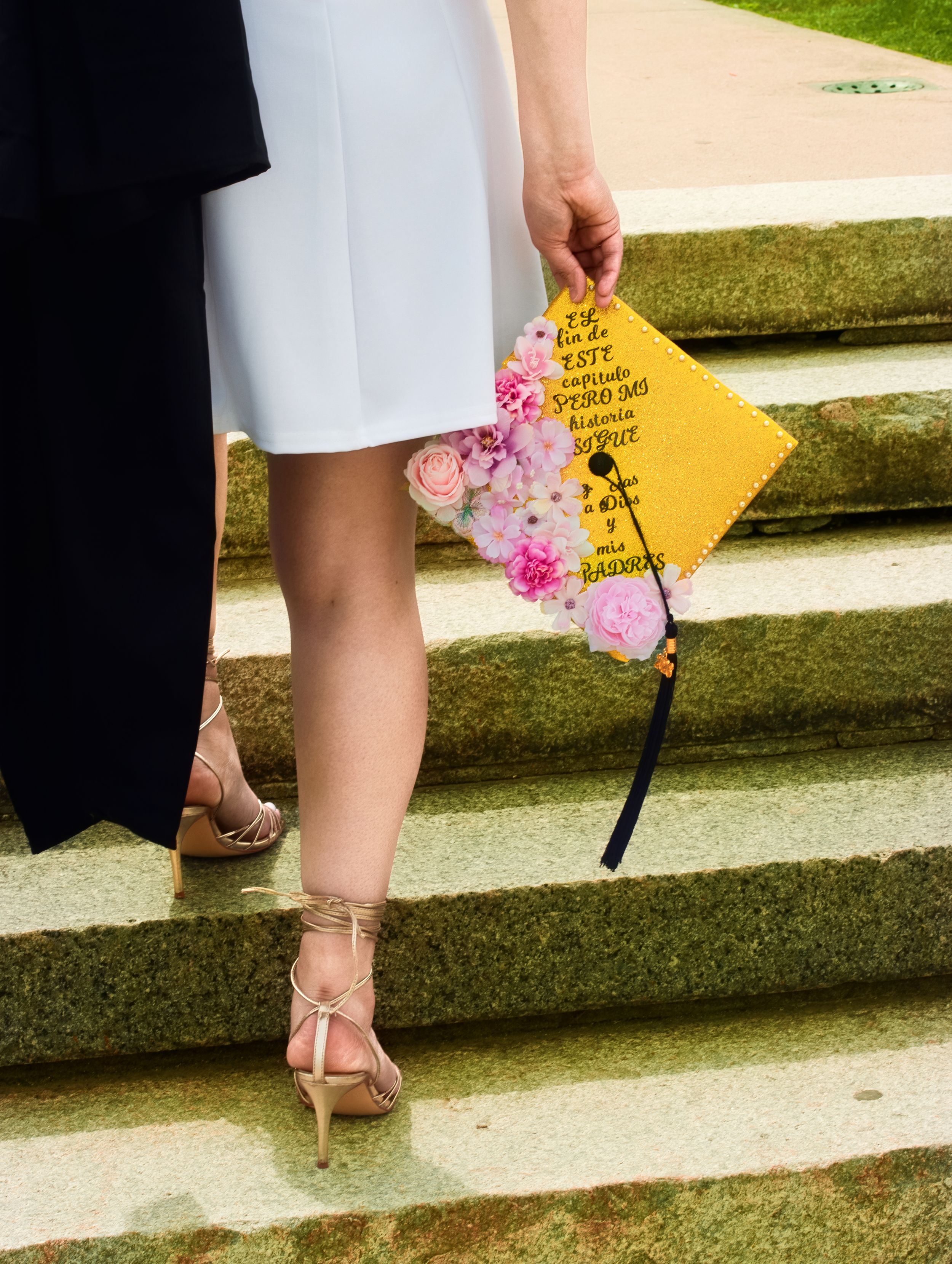 Graduate holding yellow floral mortarboard cap with Spanish text while standing on stone steps in gold heels.