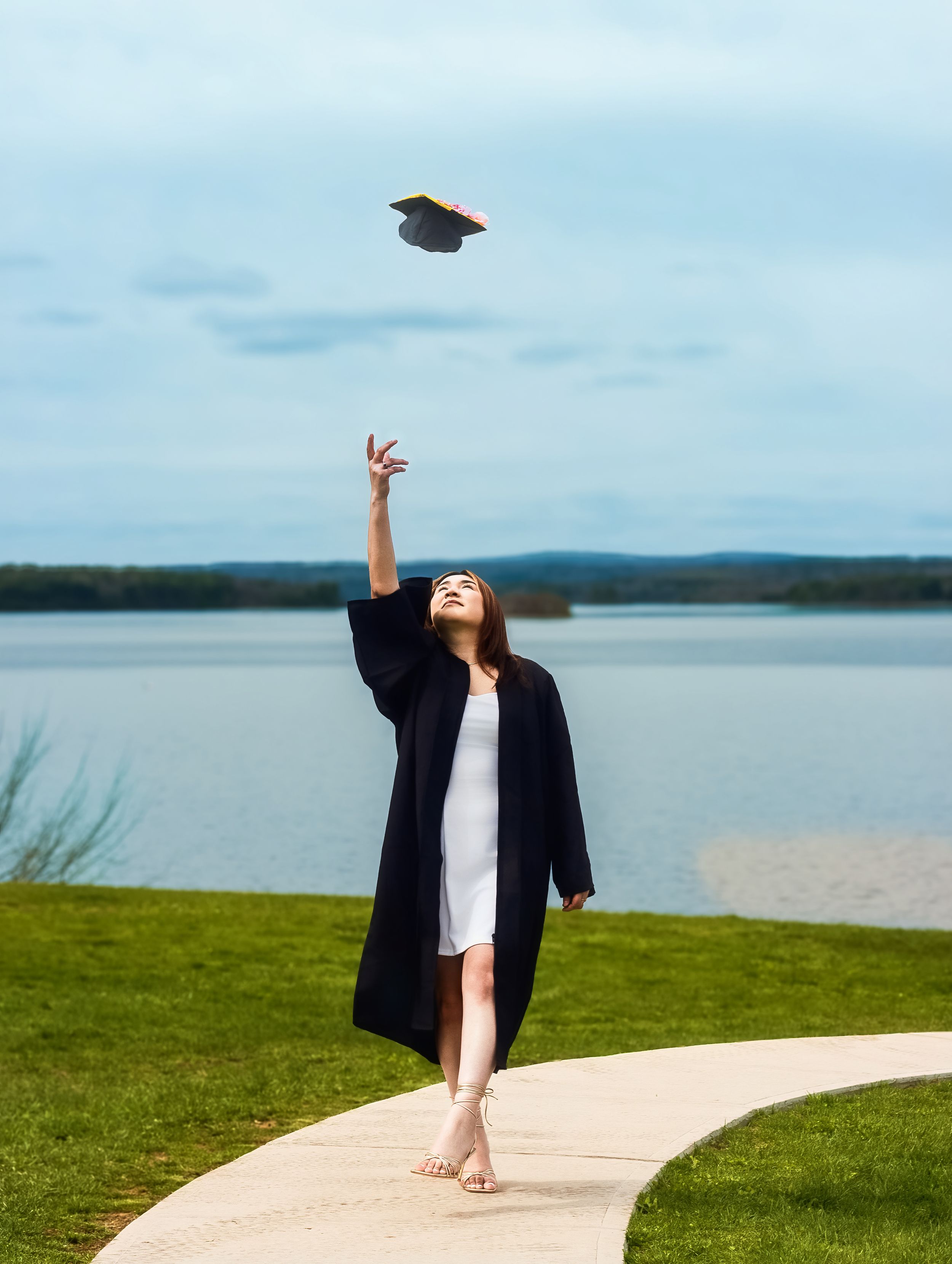 A young woman in black graduation gown tosses her cap into the air outdoors near a lake.