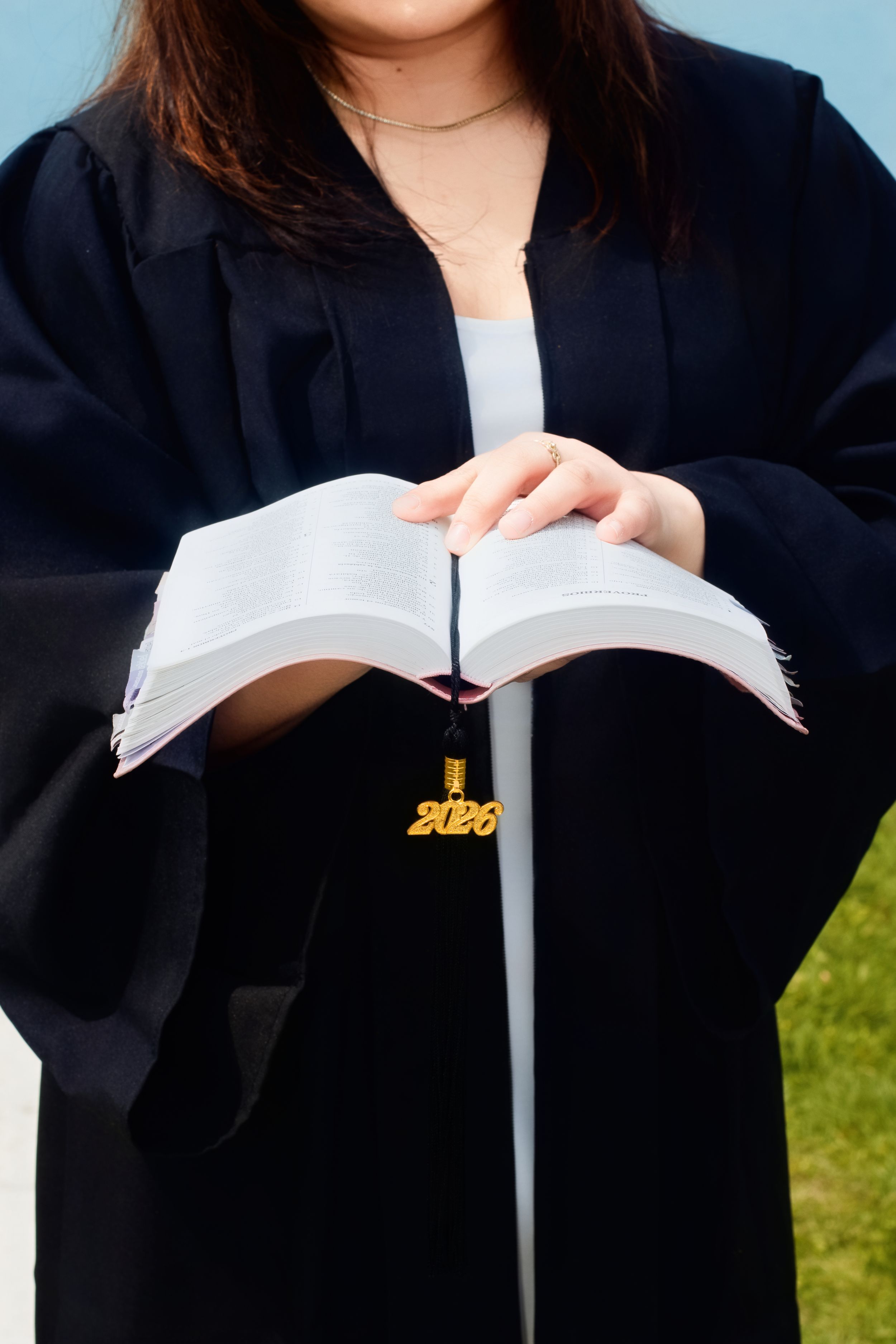 Graduate in black gown holds open book with gold 2026 tassel charm closeup.