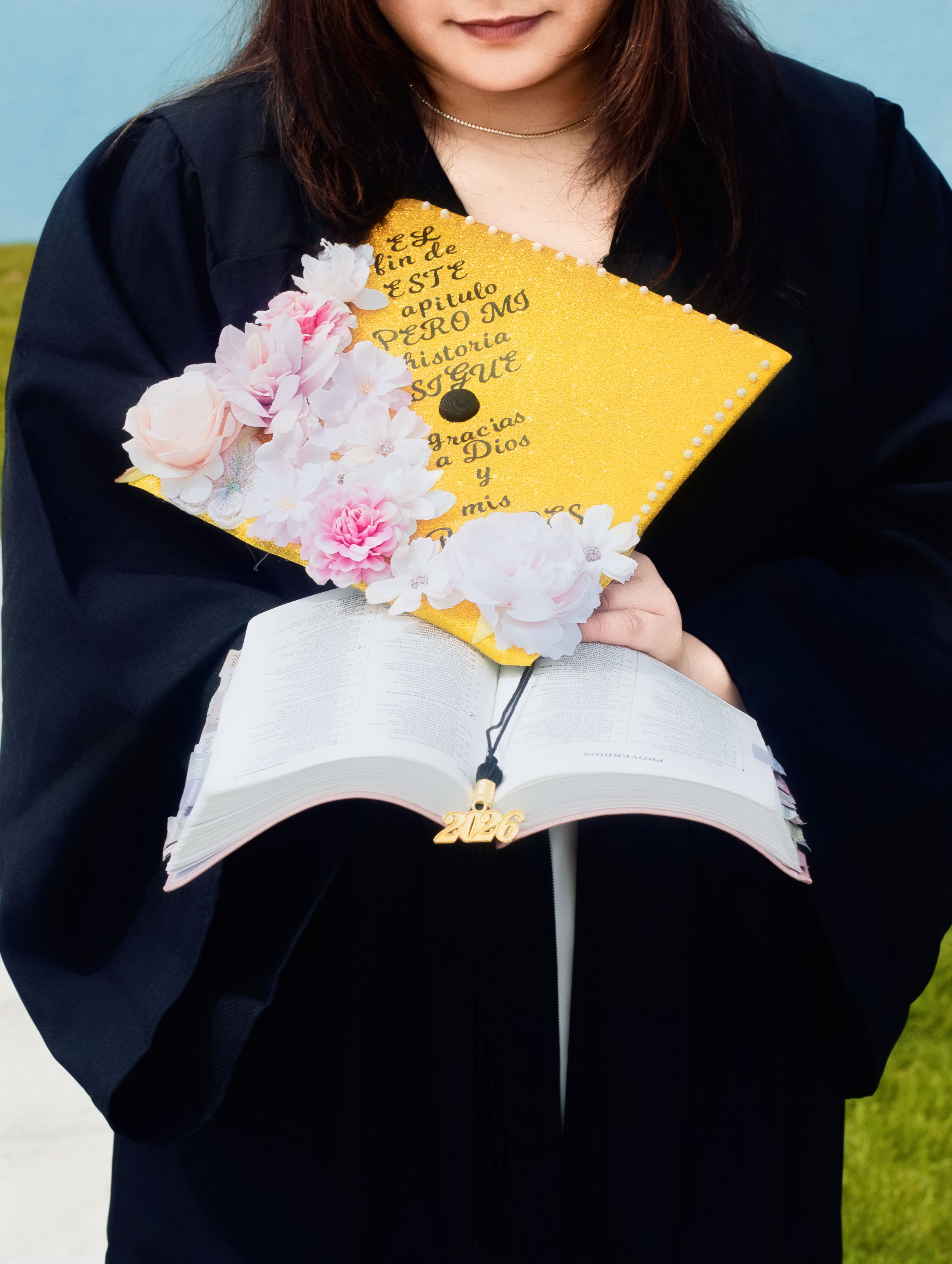 Graduate in black gown holds open Bible and decorated yellow mortarboard cap with pink flowers and Spanish text.