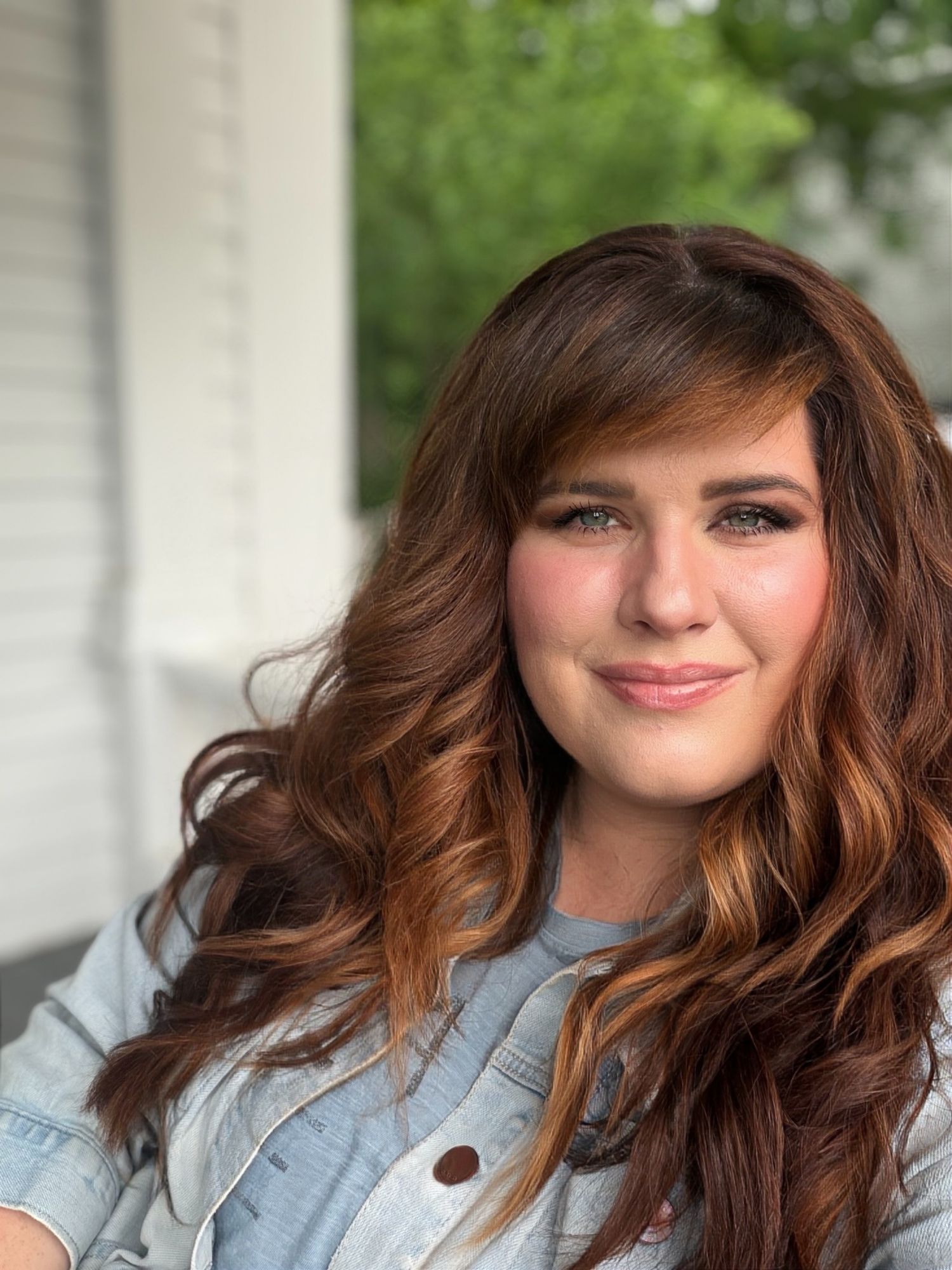 Young woman with long auburn wavy hair and bangs smiling outdoors near white porch columns.