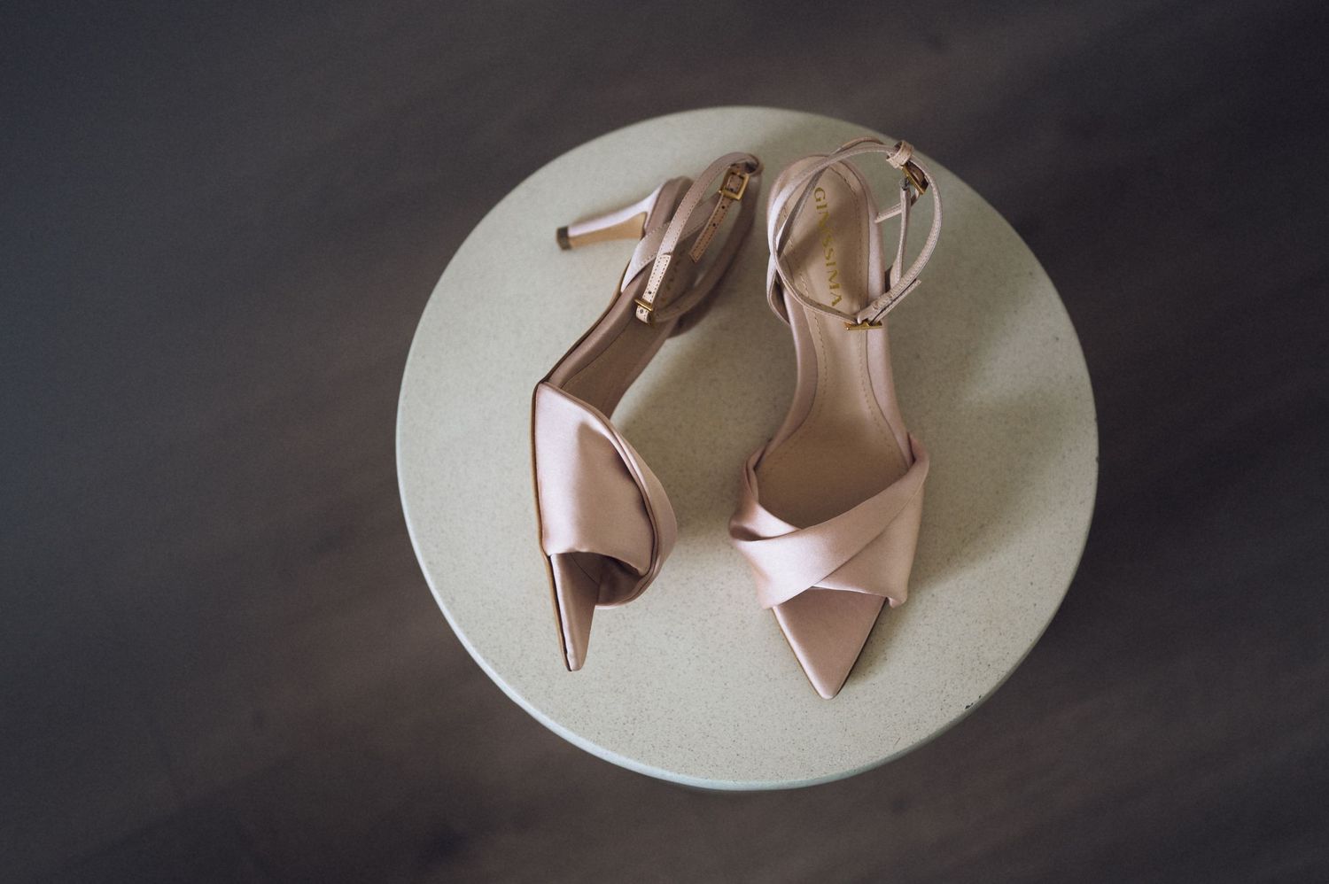 Elegant blush pink satin strappy heels with ankle strap displayed on a round cream surface.