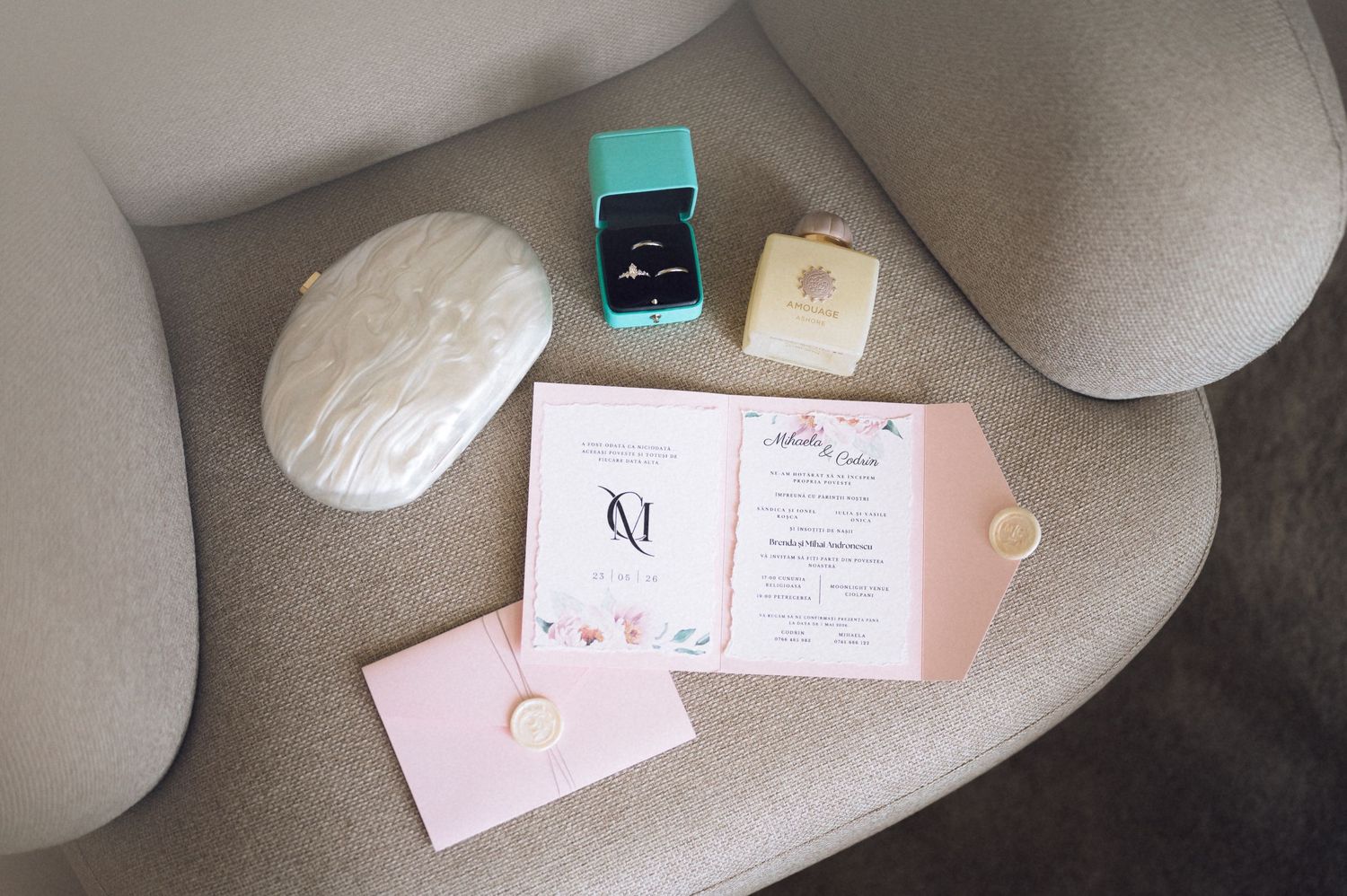 Elegant wedding flat lay with Tiffany ring box, invitation suite, and pearl clutch on a gray chair.