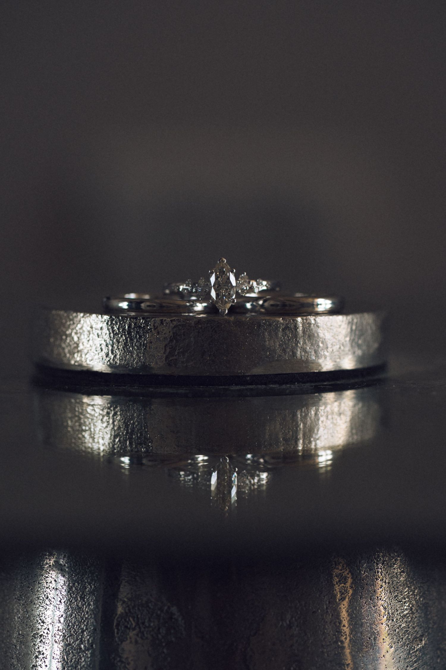 Silver wedding bands and diamond engagement ring stacked together on reflective dark surface in moody lighting.