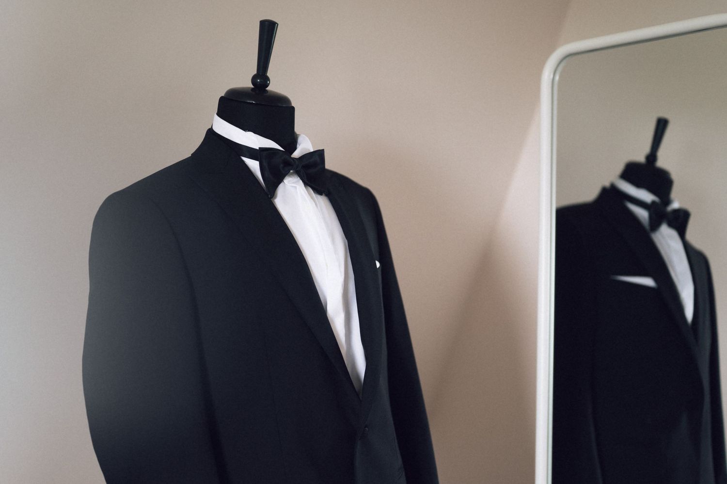 A classic black tuxedo with white dress shirt and bow tie displayed on a mannequin stand beside a mirror.